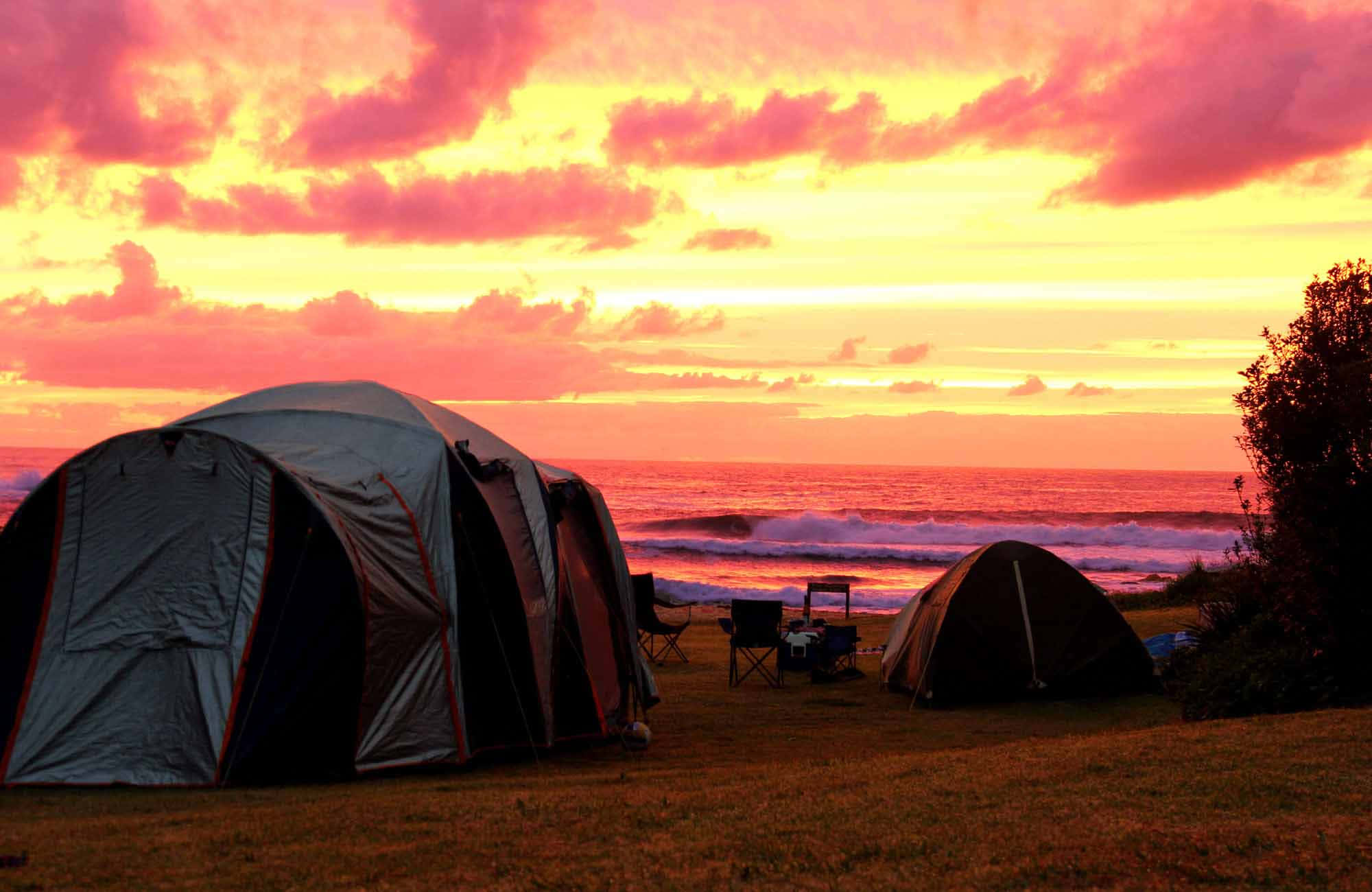 Beach Camping 桌布 壁紙 — 日落時分，草地上紮著兩個帳篷，熾熱的橙色和粉紅色雲彩在翻騰的海浪之上。