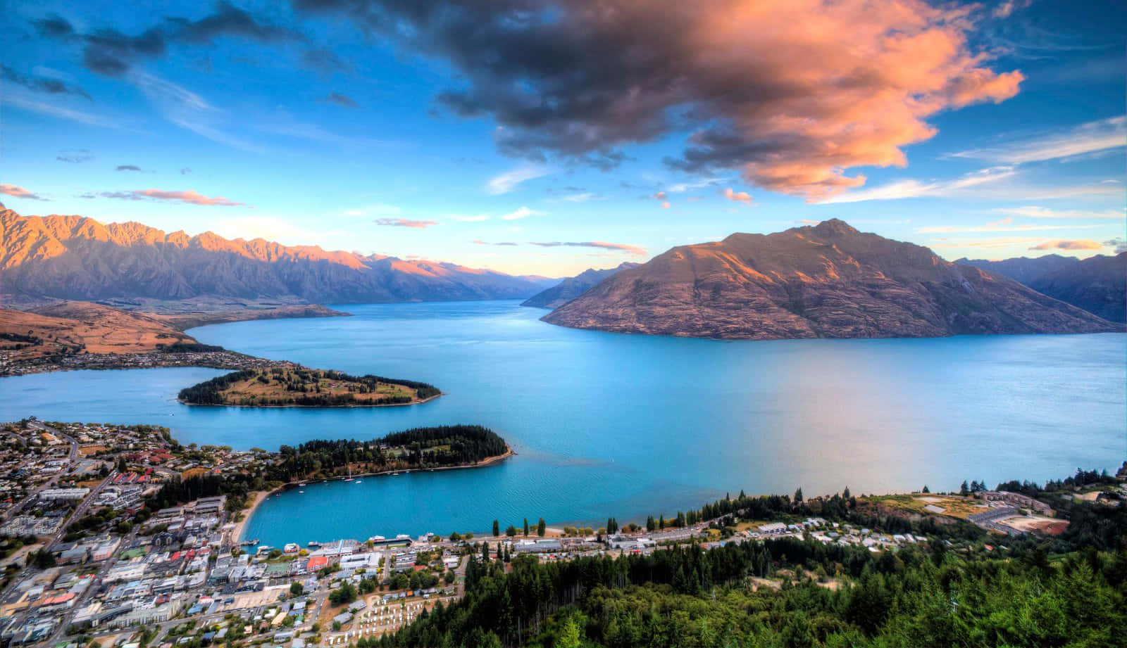 New Zealand 桌布 壁紙 — 日落時分，金色山脈之間鑲嵌著一個充滿活力的碧綠湖泊，湖岸邊有一座小鎮的鳥瞰圖。