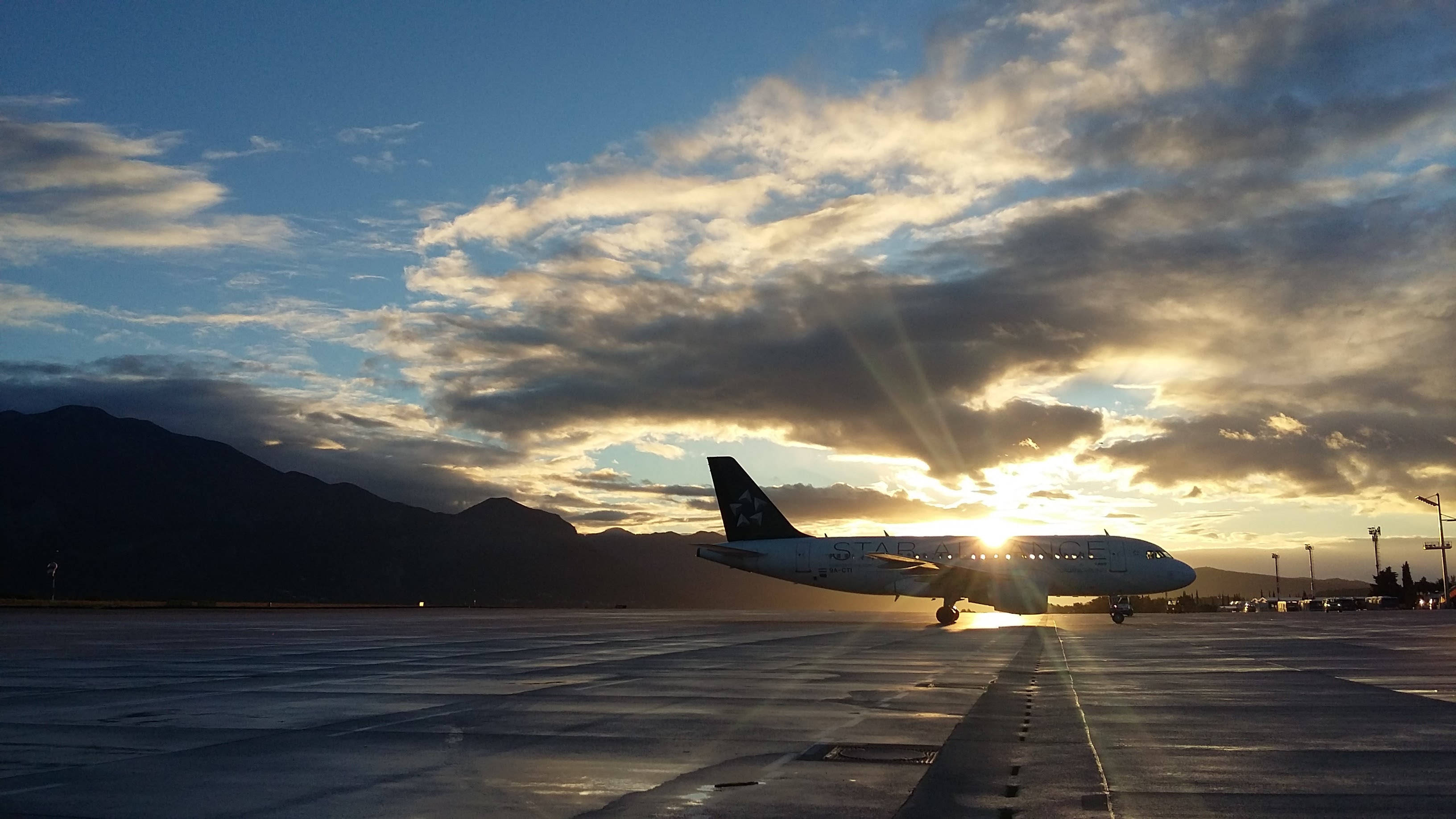 日落時分停泊飛機 壁紙 — 一架商用飛機在金色日落、戲劇性雲層和遠山背景下形成剪影。