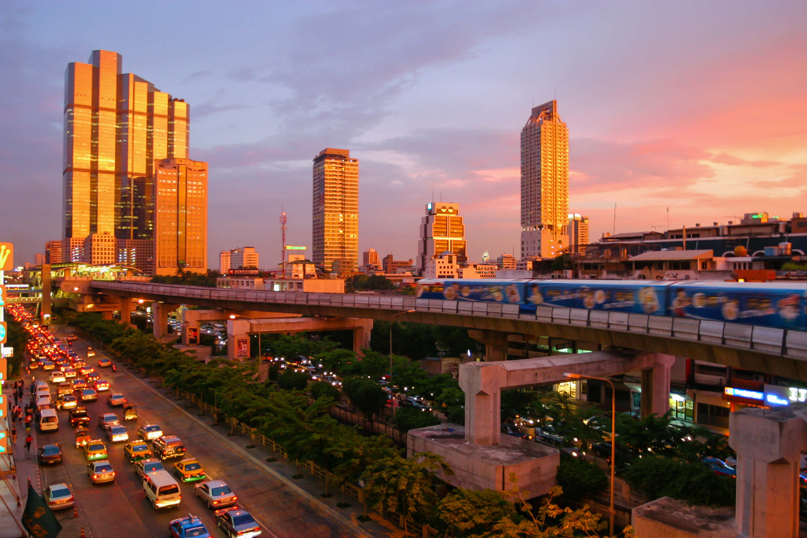 Infrastructure 桌布 壁紙 — 日落時分充滿活力的城市景觀，金色摩天大樓、繁忙的道路，以及穿梭於茂密綠樹上方的空中列車。