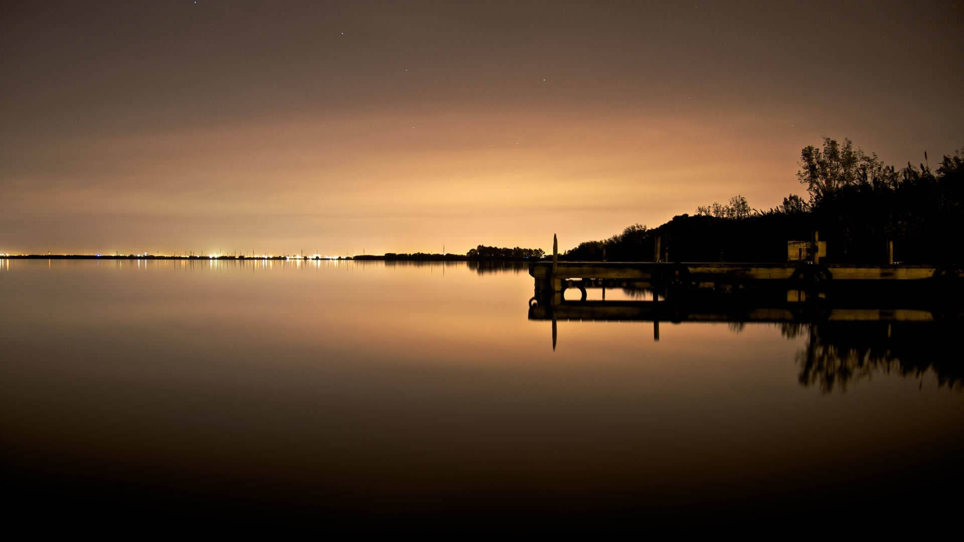 日落時分寧靜湖泊的壯麗景色 壁紙 — 寧靜的夜晚湖景，木製碼頭在發光的橙色天空映襯下形成剪影，倒映在平靜的水面上。