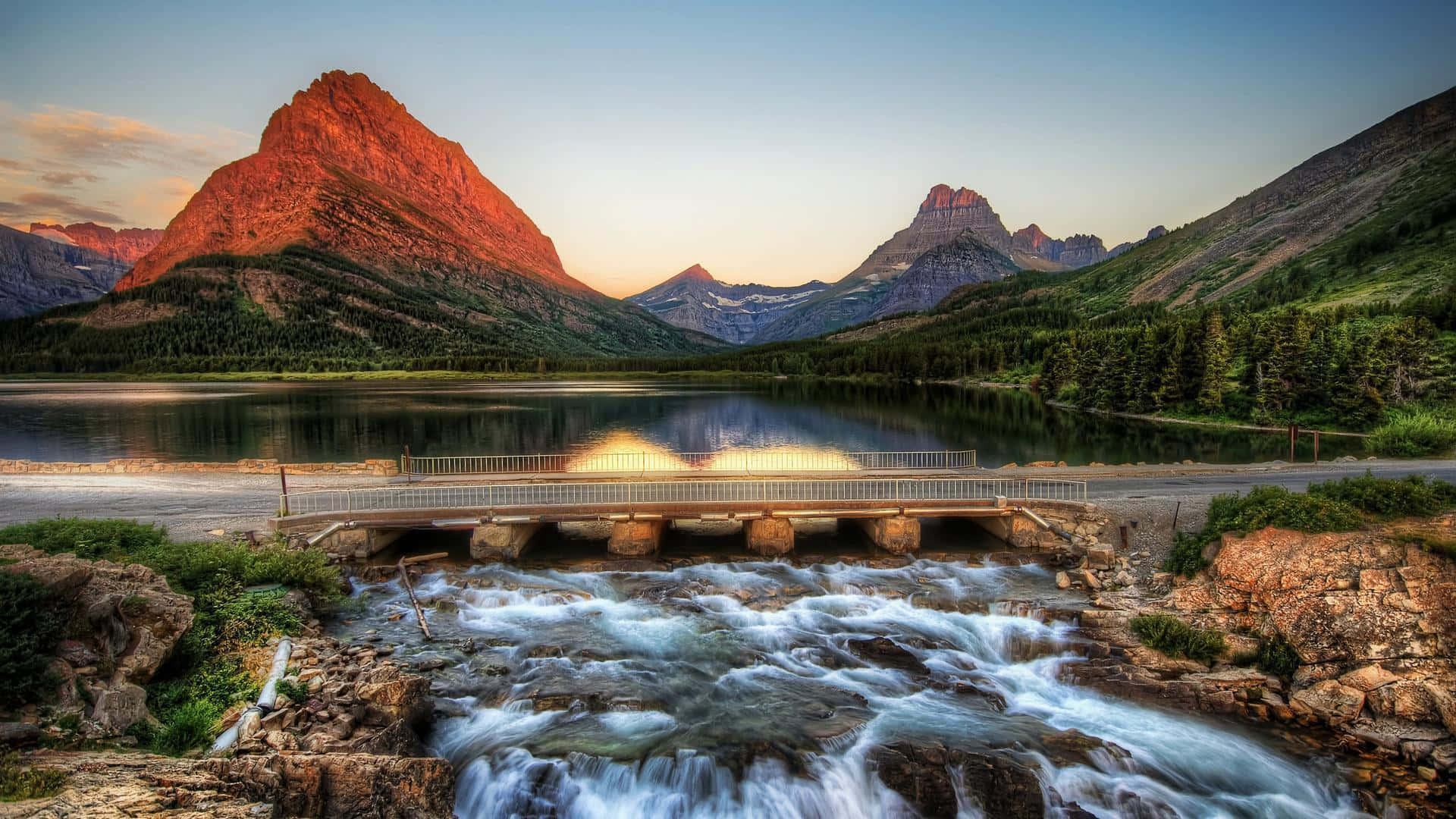 Montana 桌布 壁紙 — 日落時分寧靜的山間湖泊，一座橋樑橫跨湍急的水流，白雪覆蓋的山峰倒映在平靜的水面上。