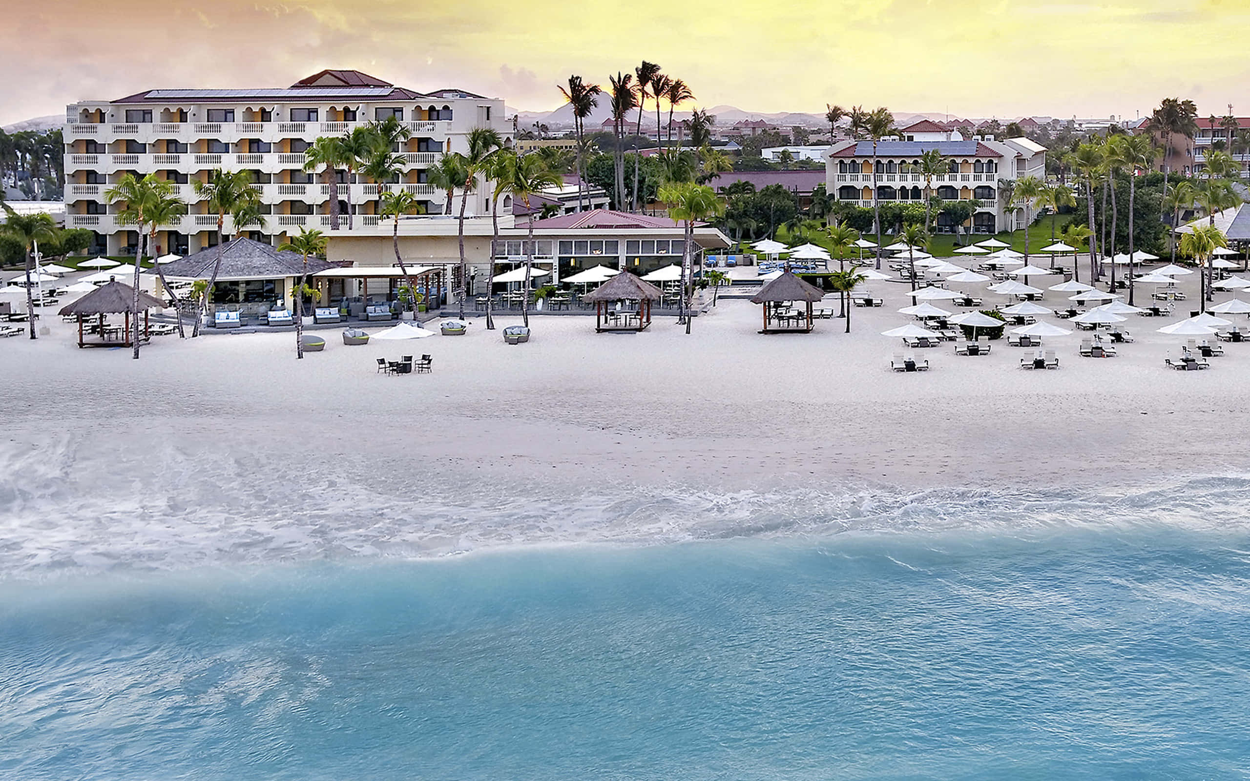 Beach Resort 桌布 壁紙 — 日落時分寧靜的熱帶海灘，有白色沙灘、碧綠海水和棕櫚樹環繞的度假村建築。