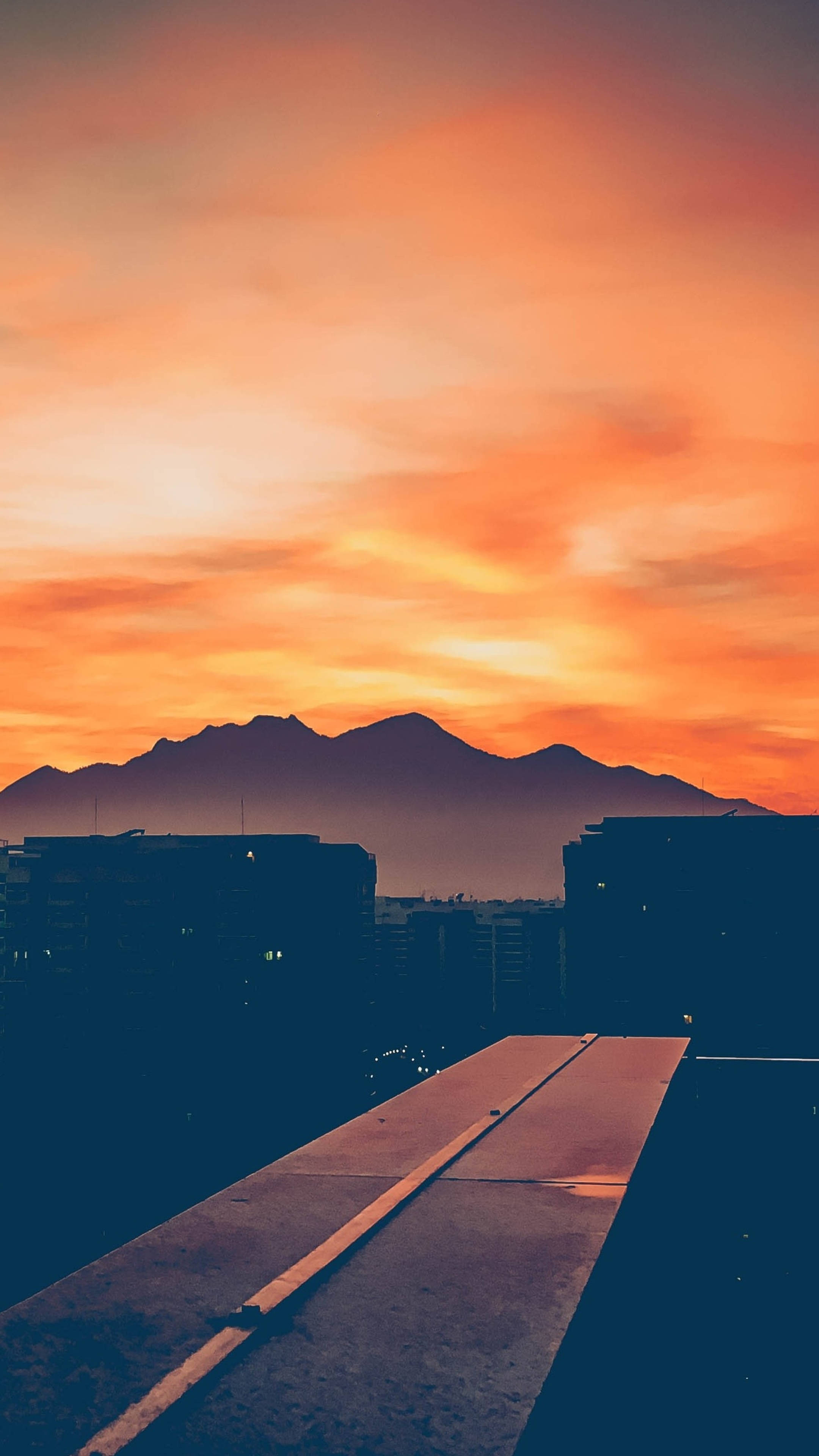 Sunset iPhone 桌布 壁紙 — 日落時分從屋頂俯瞰，火紅橙色的雲層籠罩在黑暗剪影般的山巒和城市建築之上。