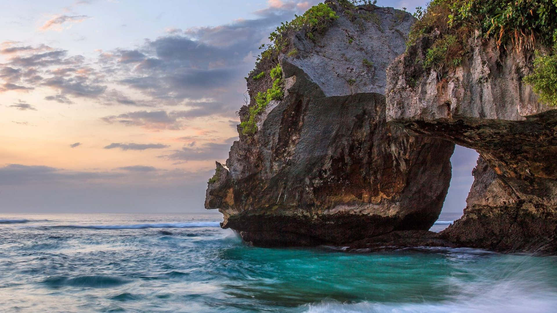 Bali Beach 桌布 壁紙 — 日落時分戲劇性的岩石拱門，下方有碧綠色的海水拍打，上方有柔和的粉紅色雲彩。