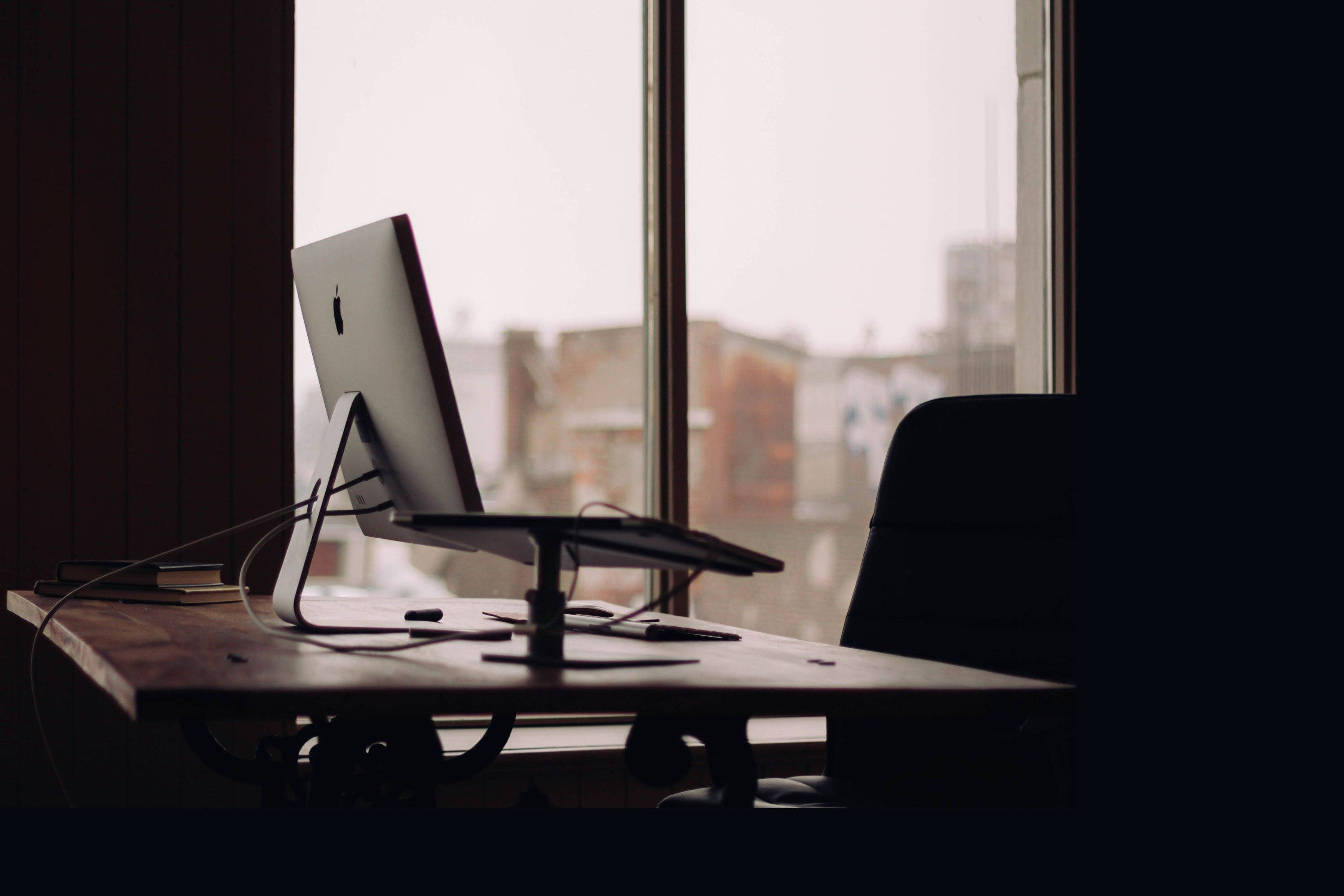 日落時分木質辦公桌 壁紙 — 一部時尚的 iMac 放在木桌上，旁邊是窗戶，窗外是柔和日光下的朦朧城市景觀。