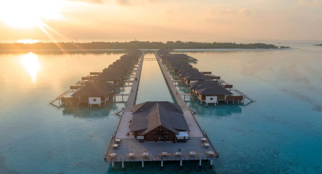 Maldives Island 桌布 壁紙 — 日落時分水上平房的鳥瞰圖，金色光線反射在碧綠的水面上。