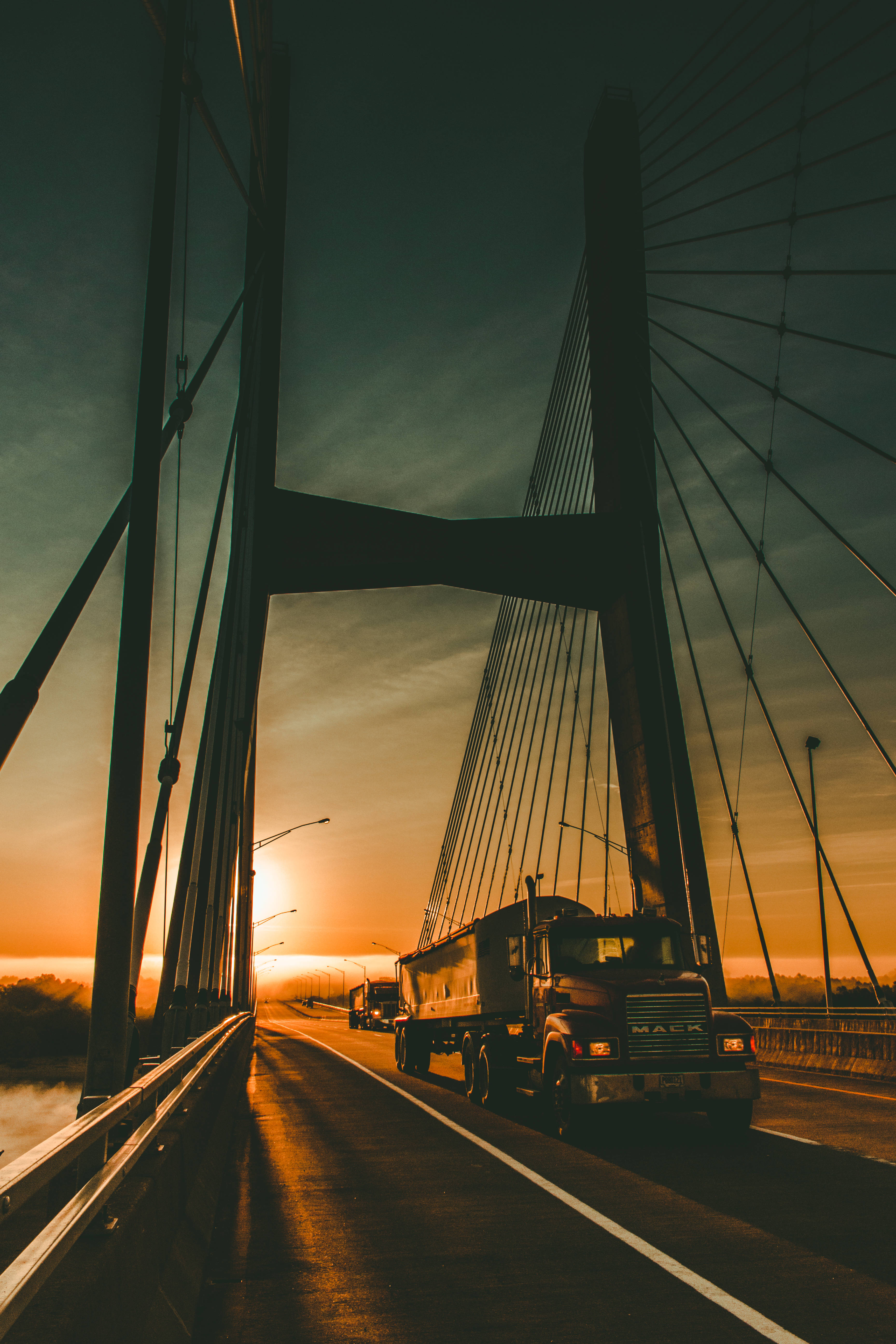 日落時分的卡佩吉拉德橋貨車 壁紙 — 一輛巨型卡車駛過斜拉橋，夕陽在它後面落下，將金色光芒灑在路上。