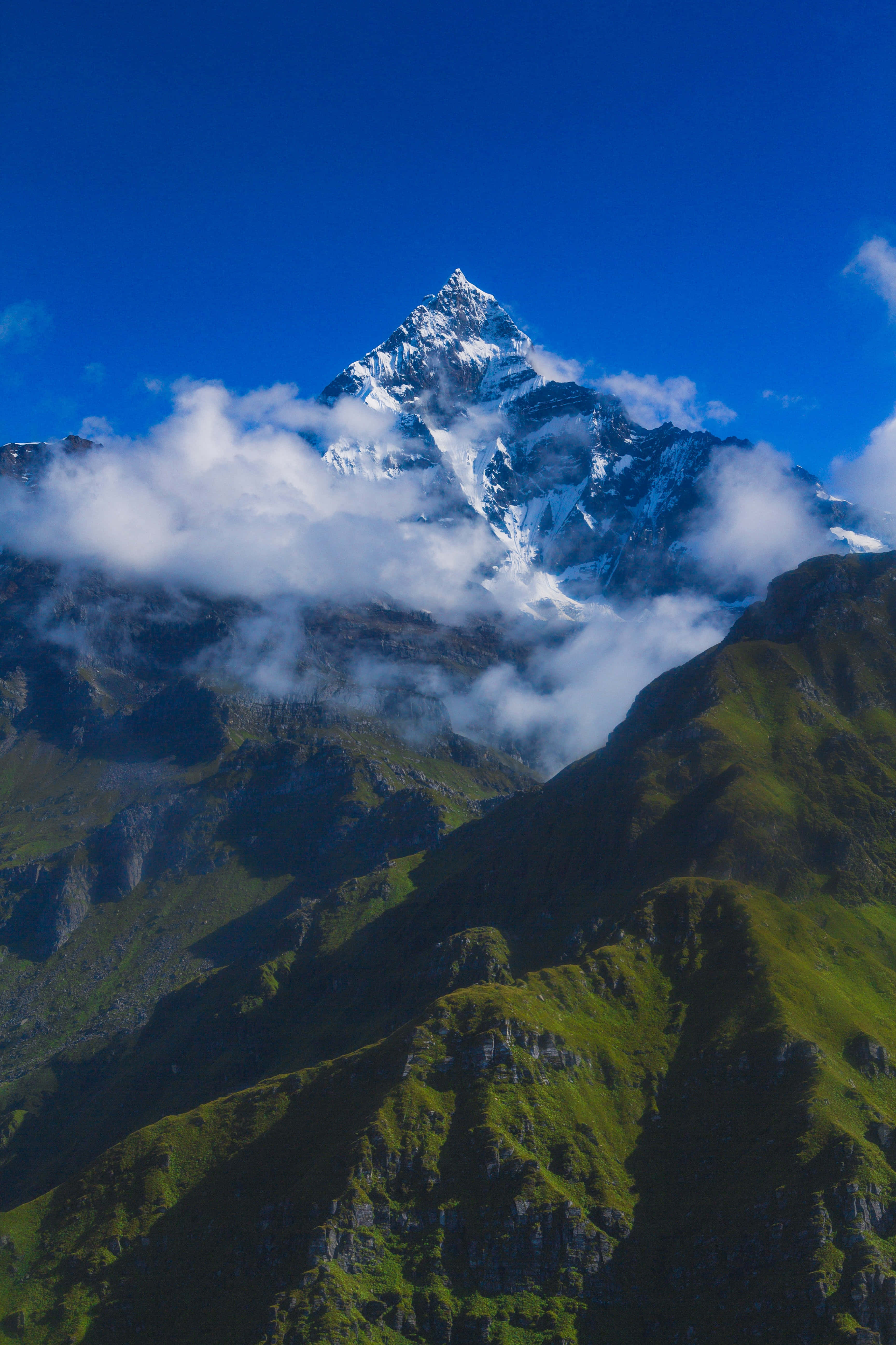 日落時分的壯麗山景 壁紙 — 雄偉的雪山峰穿過蓬鬆的白雲，映襯著充滿活力的藍天，下方是鬱鬱蔥蔥的綠色山坡。