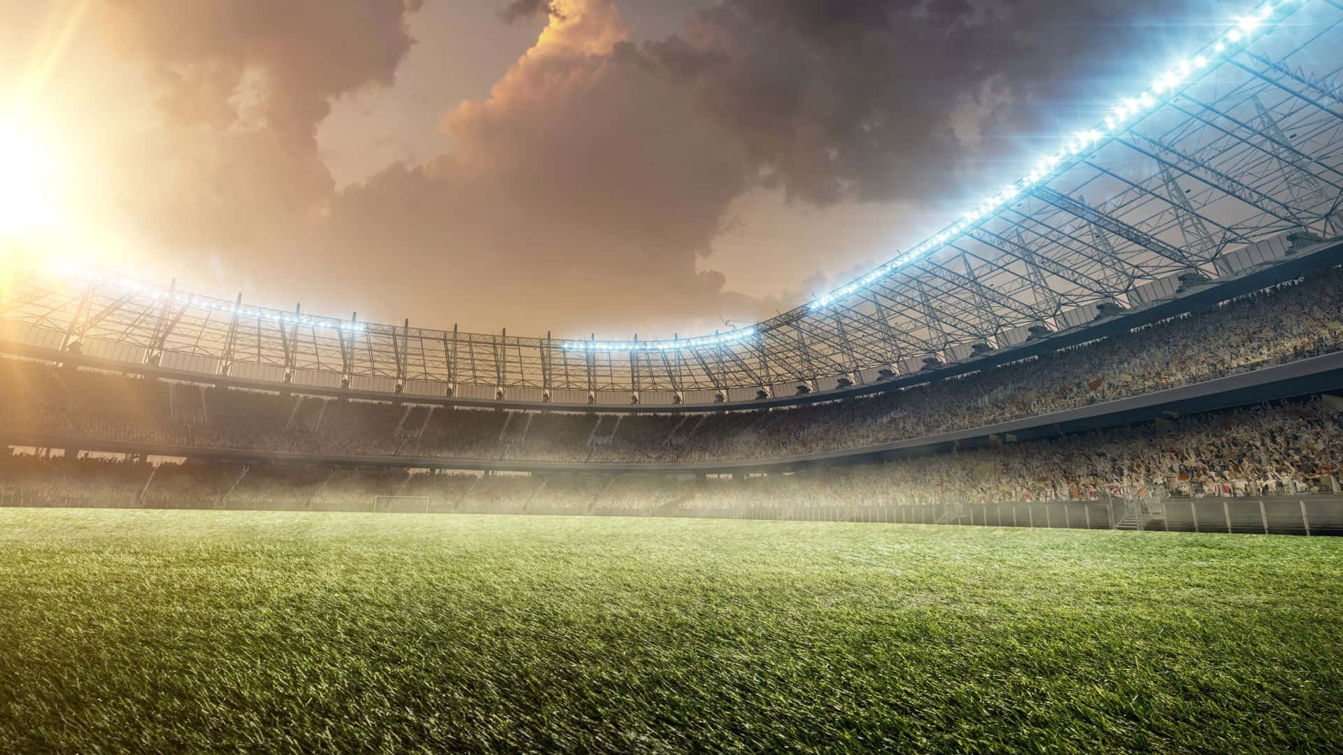 Soccer Stadium 桌布 壁紙 — 日落時分的廣闊體育場，戲劇性的雲層下，明亮的泛光燈照亮了翠綠的草地和座無虛席的看台。