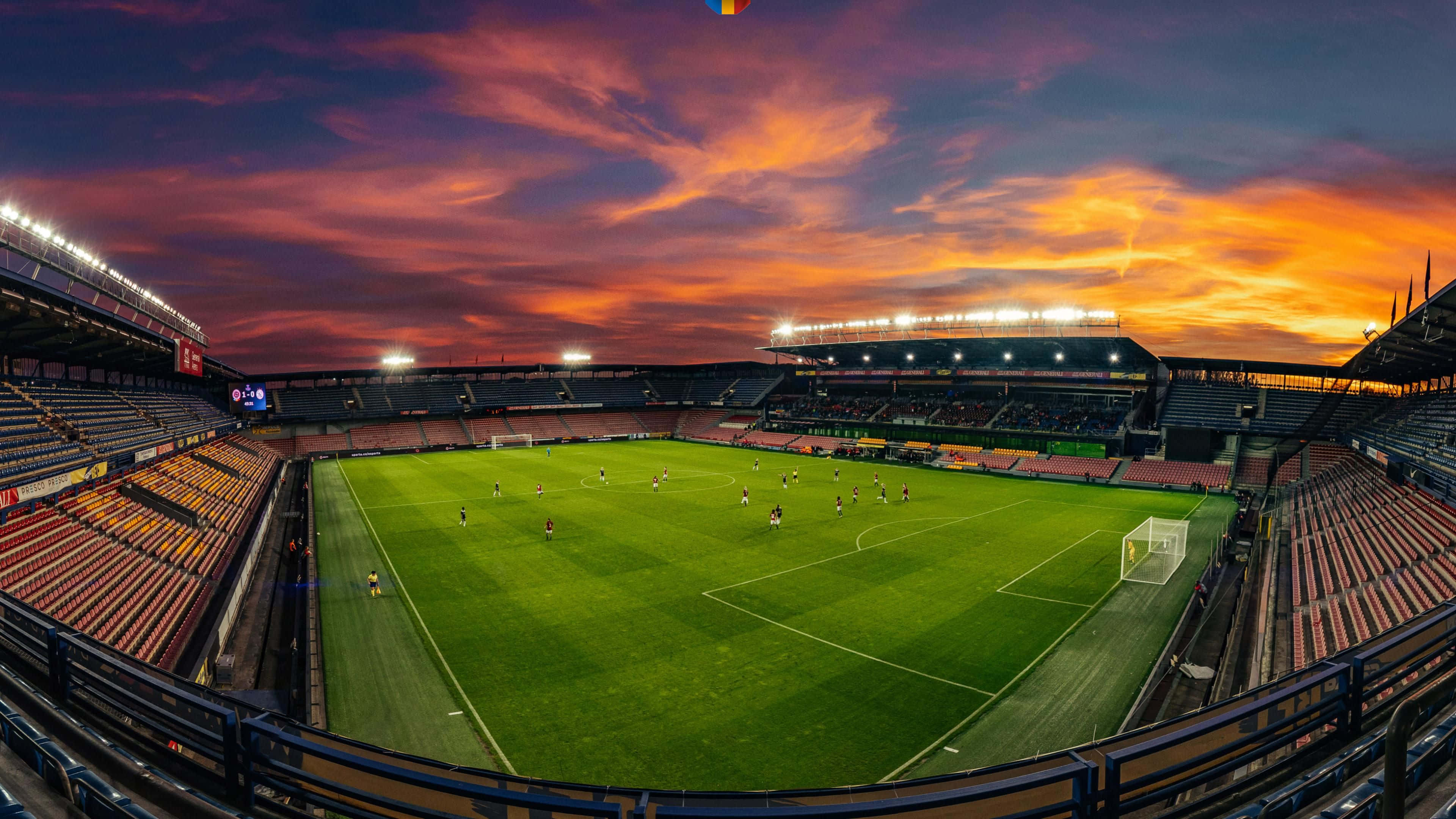 Soccer Stadium 桌布 壁紙 — 日落時分的足球場，場上有球員，上方有鮮豔的橙粉紅雲彩。