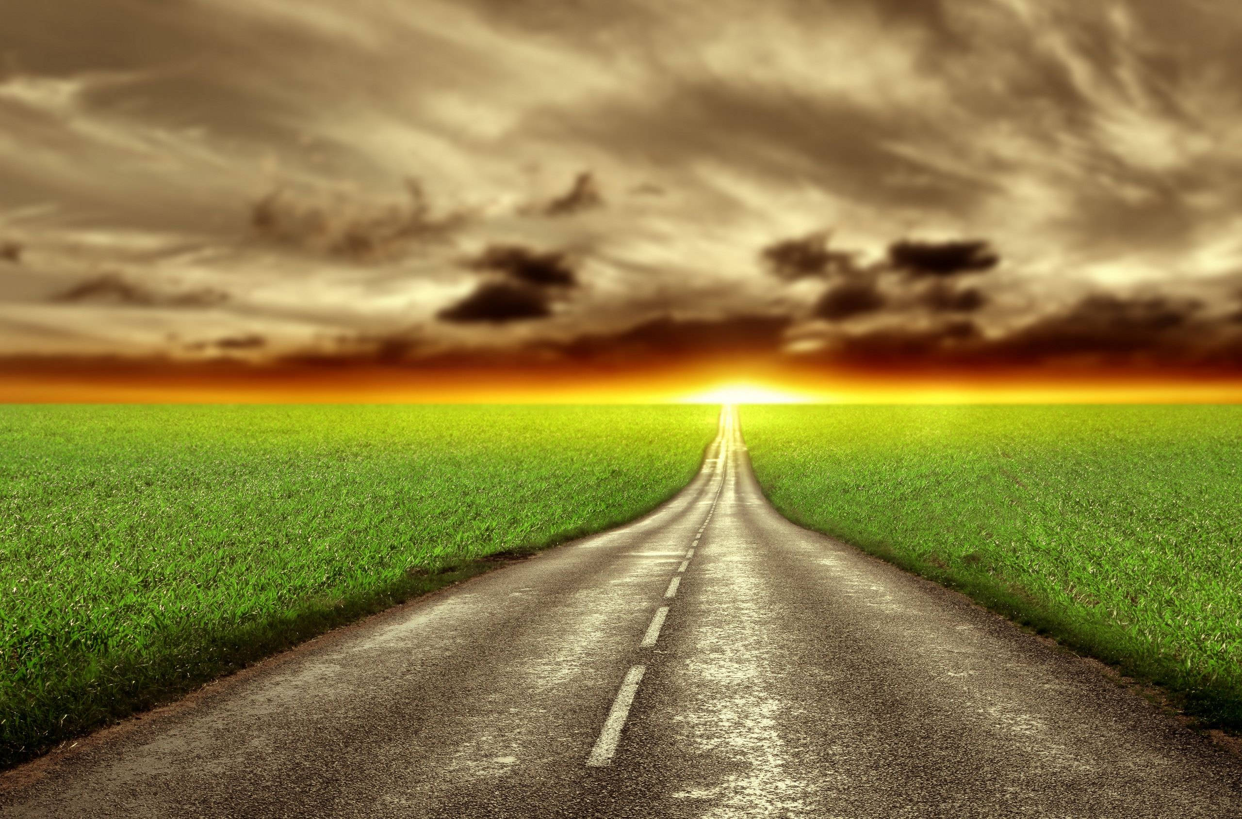 日落時分的鄉村道路 壁紙 — 一條蜿蜒的瀝青路穿過充滿活力的綠色田野，朝向戲劇性雲層下閃耀的日落地平線。