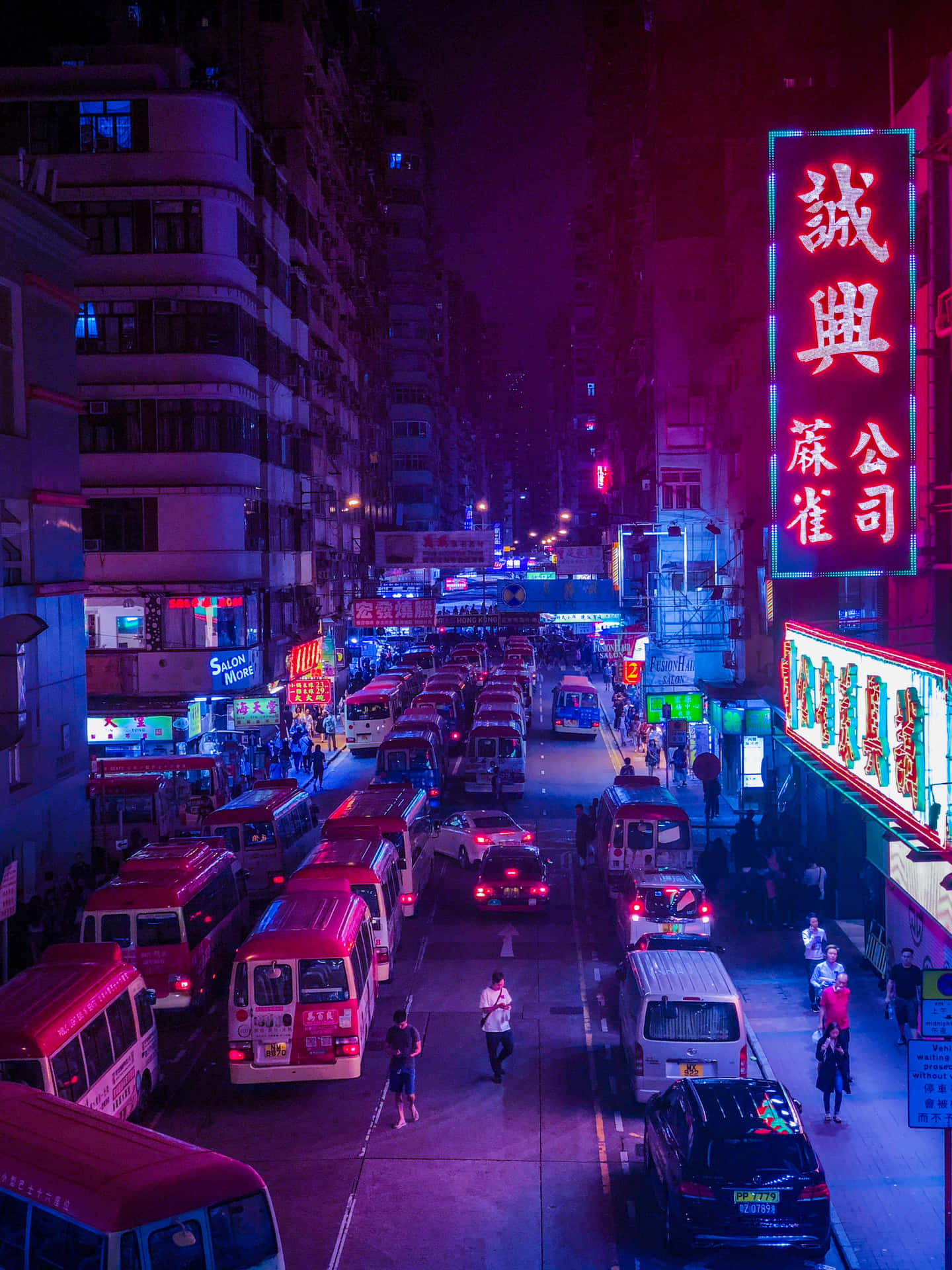 日落時分的香港城市天際線 壁紙 — 夜晚一條被霓虹燈浸透的城市街道，紅色巴士和發光招牌在擁擠的人行道上投射出鮮豔的紫色和粉紅色光芒。