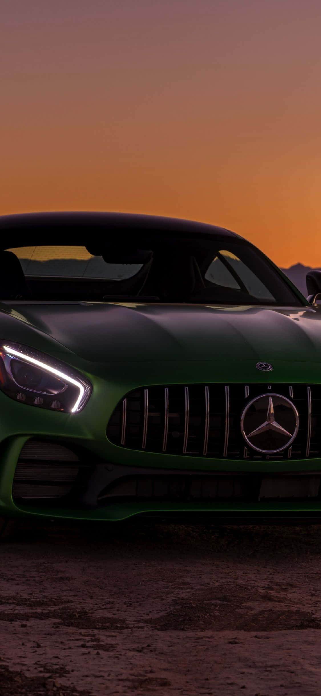 日落時分綠色iPhone XS與梅賽德斯-AMG GT背景 壁紙 — 一輛時尚的綠色 Mercedes-AMG 跑車在溫暖的橙色日落天空下閃閃發光。