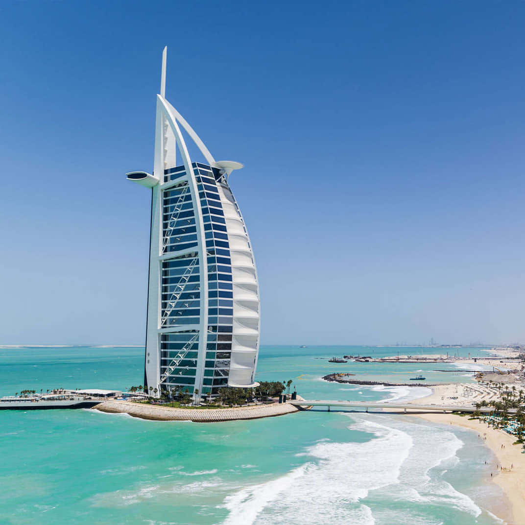 日落時分迪拜帆船酒店的迷人風景 壁紙 — 帆船形的帆船酒店（Burj Al Arab Jumeirah）雄偉地矗立在人工島上，在清澈的藍天下映襯著碧綠的海水。