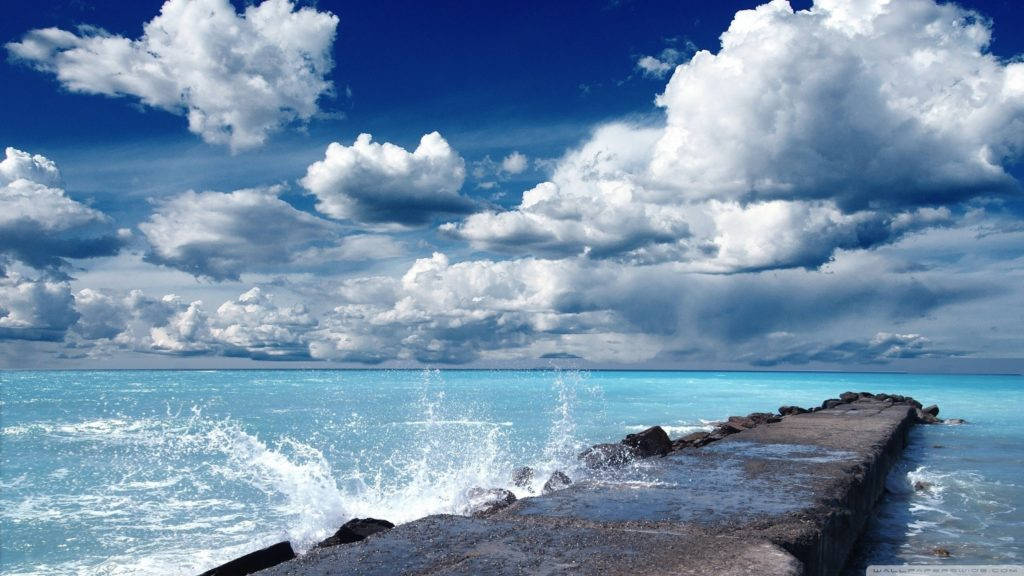 日落時分雄偉山脈的寧靜美景 壁紙 — 強烈的藍綠色海浪拍打著風化的石頭碼頭，在廣闊的天空下，天空佈滿了戲劇性的白雲。
