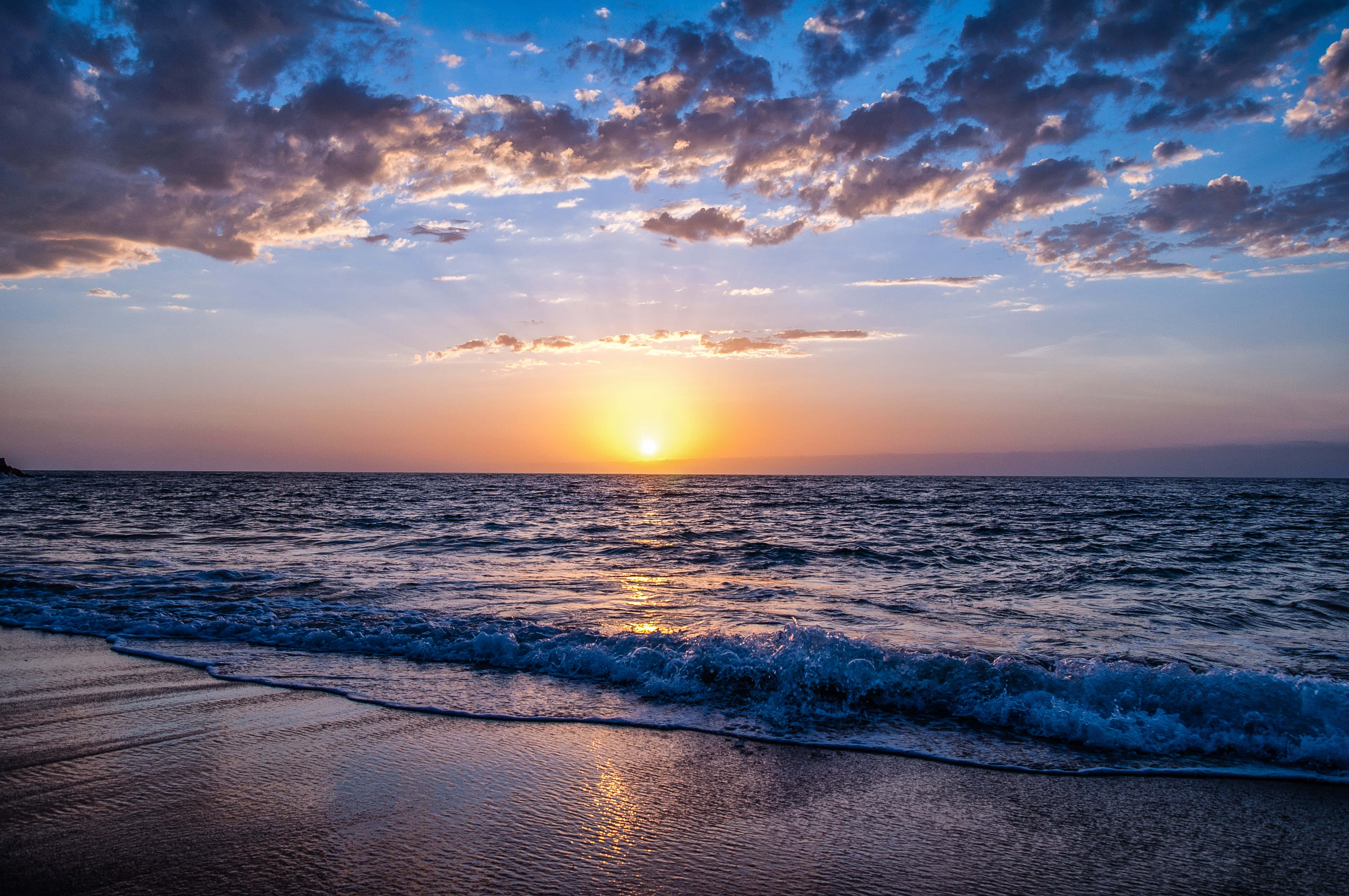 Sunset Ocean 桌布 壁紙 — 日落海洋，金色光芒映照在波浪上，拍打濕沙，天空有紫色和橙色雲彩。