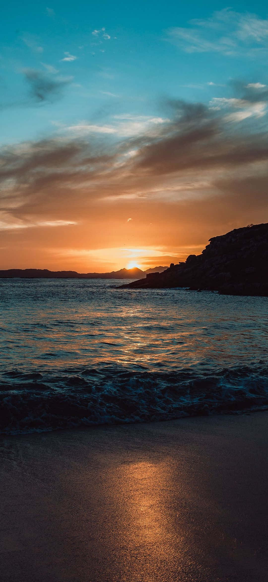 日落海灘手機背景 壁紙 — 夕陽將天空染成火橙色和青綠色，海浪輕輕拍打岩石懸崖旁的沙灘。