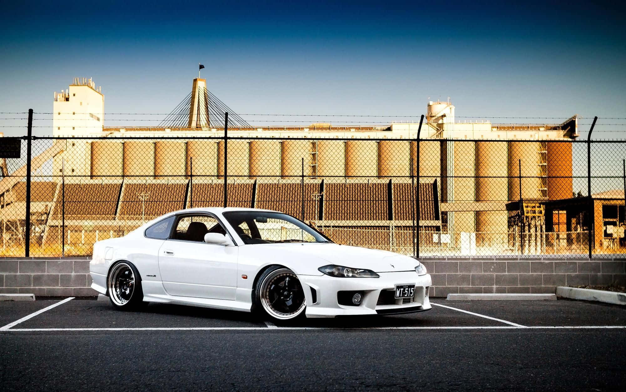 日製尼桑Silvia S15 壁紙 — 一架純白色跑車停泊在空地上，鐵絲網後面有工業建築物，天空清澈湛藍。