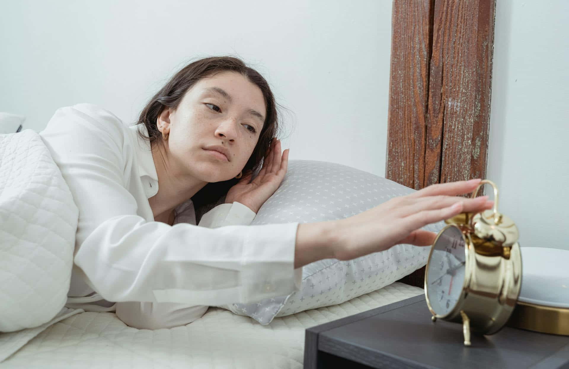 早晨鬧鐘例行程序 壁紙 — 一位穿著白襯衫的疲倦女子躺在床上，枕著枕頭，伸手去拿金色的鬧鐘。