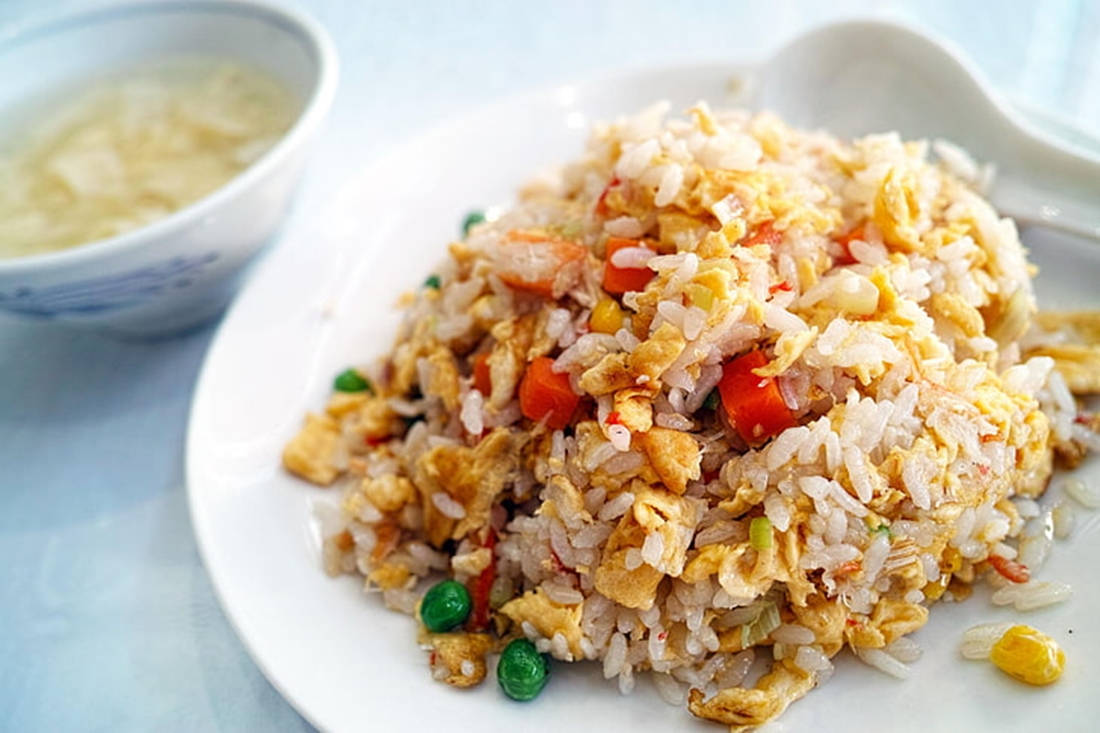 早餐炒飯 壁紙 — 一個特寫鏡頭，顯示一堆色彩繽紛的炒飯，配有雞蛋、蔬菜和玉米，放在白色碟子上，旁邊有一碗湯。