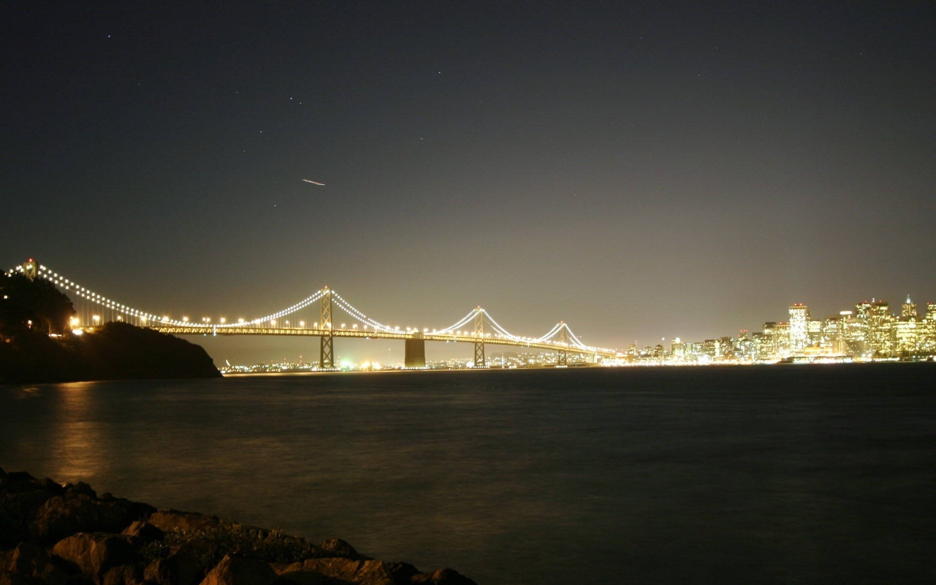 星空下，夜間城市燈光映照水面上的一座亮燈吊橋。