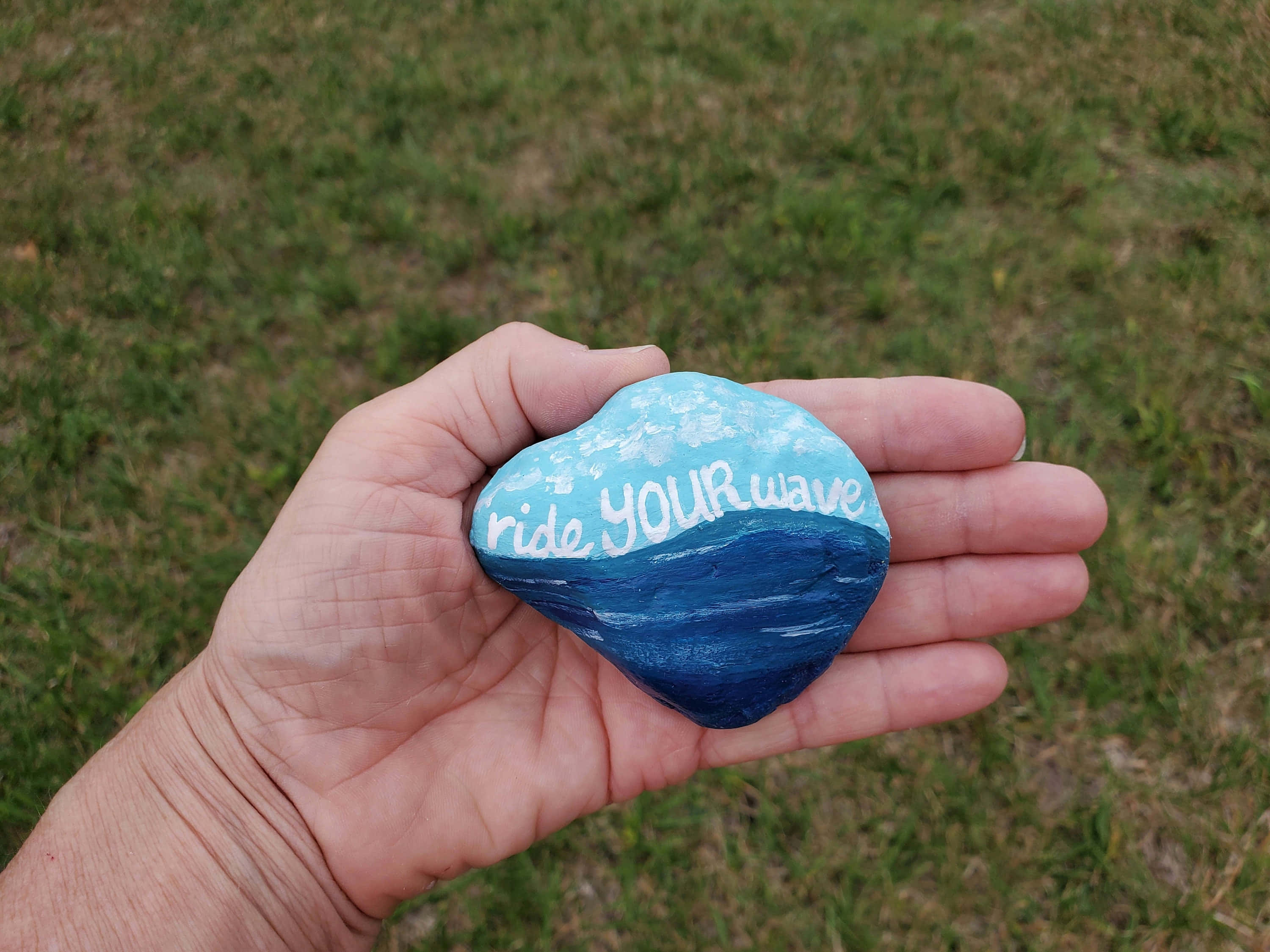 明亮多彩的 Pebble Art 壁紙 — 一隻手拿著一塊畫有海浪圖案的石頭，上面用白色草書寫著「ride YOUR wave」，背景是草地。