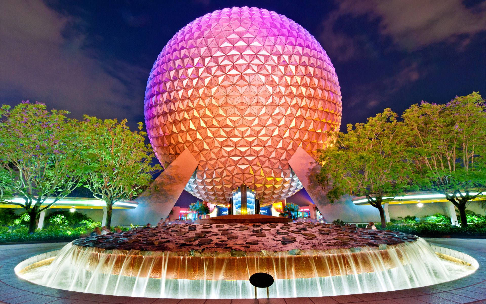 明亮橙色 Epcot 球體 壁紙 — 一個巨大的測地線球體在夜間發出紫色和橙色光芒，位於一個圓形噴泉上方，周圍環繞著樹木。
