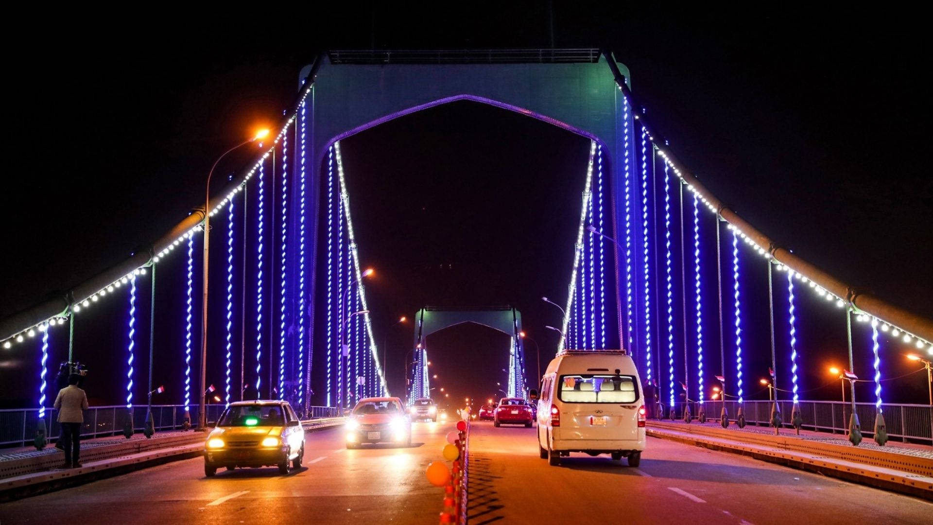 明亮的巴格達橋 壁紙 — 現代吊橋上充滿活力的夜景，橋上藍白燈光如瀑布般閃爍，汽車在其發光的拱門下行駛。