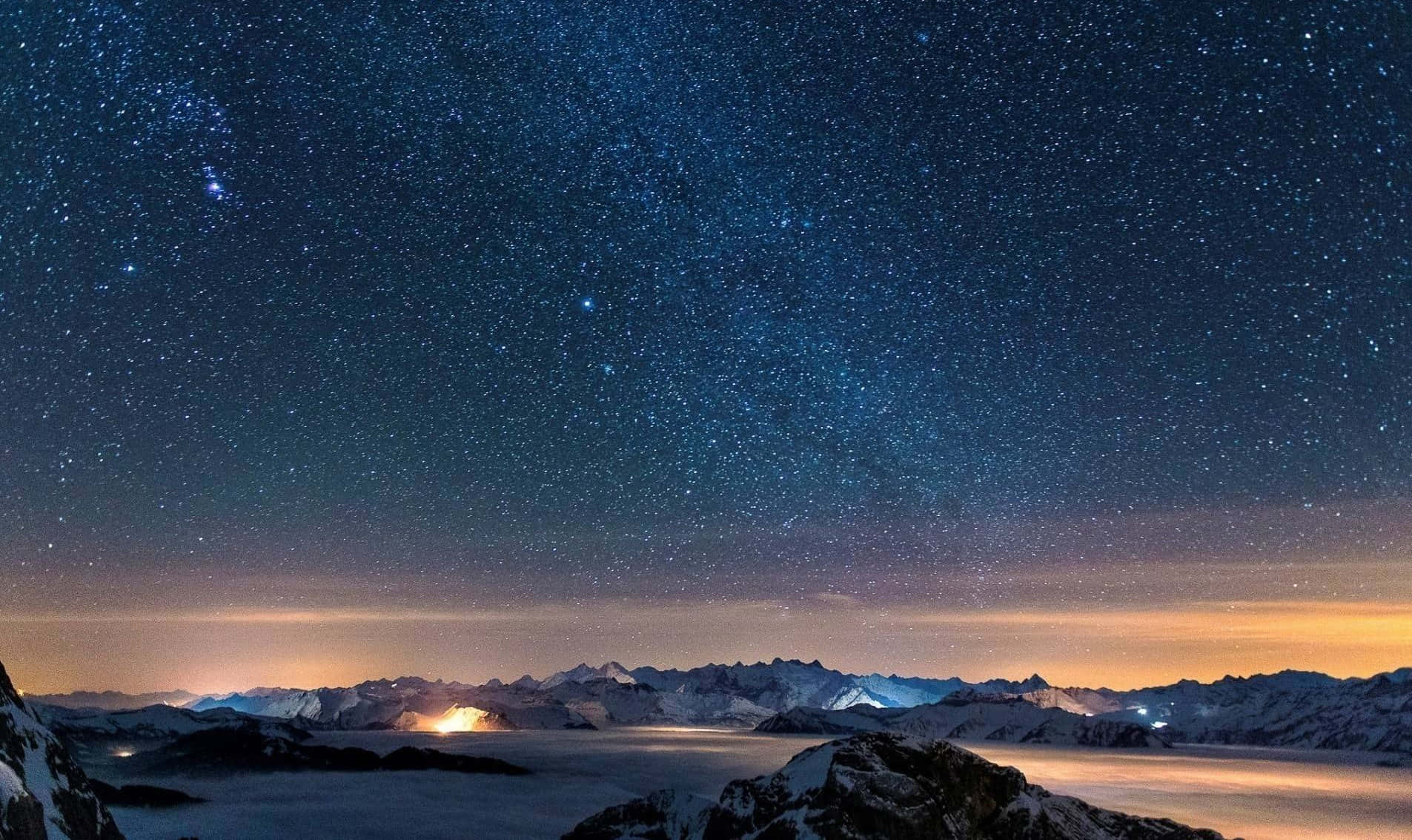 明亮的星星照亮充滿希望的夜空 壁紙 — 在繁星點點的夜空下，積雪的山峰伴隨著發光的極光和遠處的燈光。