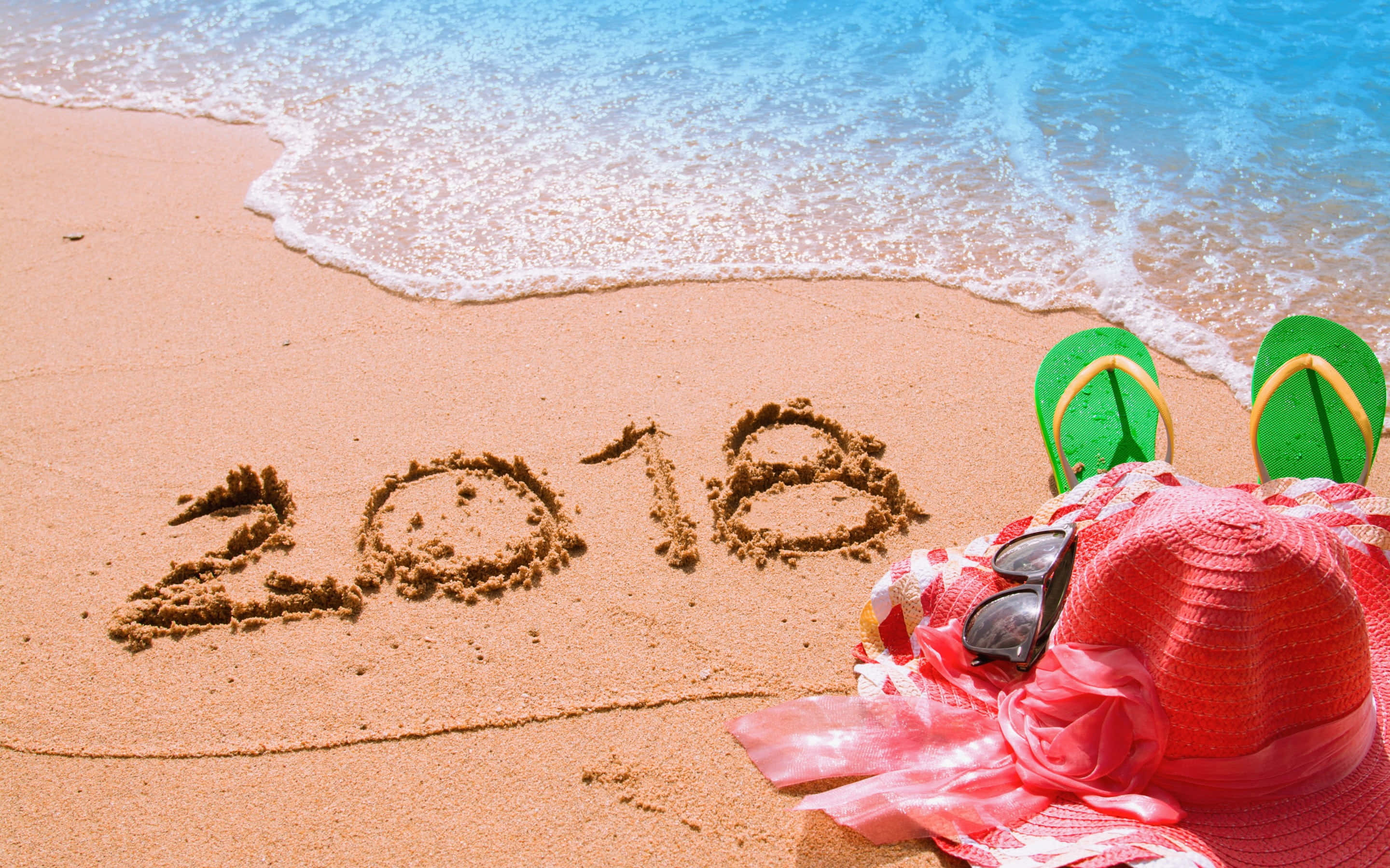 Beach Accessories 桌布 壁紙 — 明亮的沙灘場景，沙灘上寫著「2018」，旁邊有粉紅色帽子、太陽眼鏡和綠色人字拖，海浪翻滾而來。
