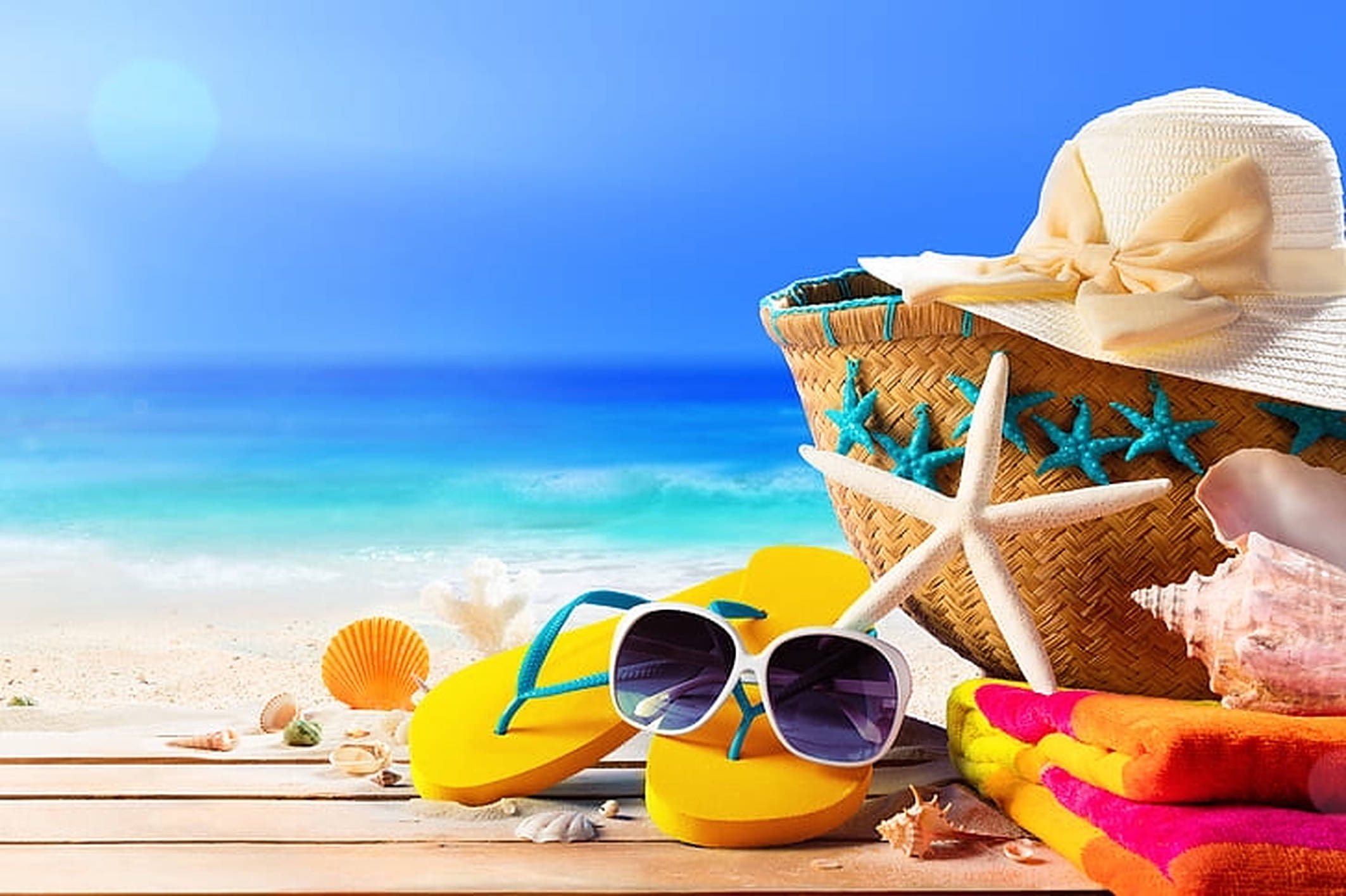 Beach Vacation 桌布 壁紙 — 明亮的沙灘配件，包括黃色人字拖、太陽眼鏡、海星和貝殼，放在木板上，旁邊是碧綠色的海洋，上方是清澈的藍天。