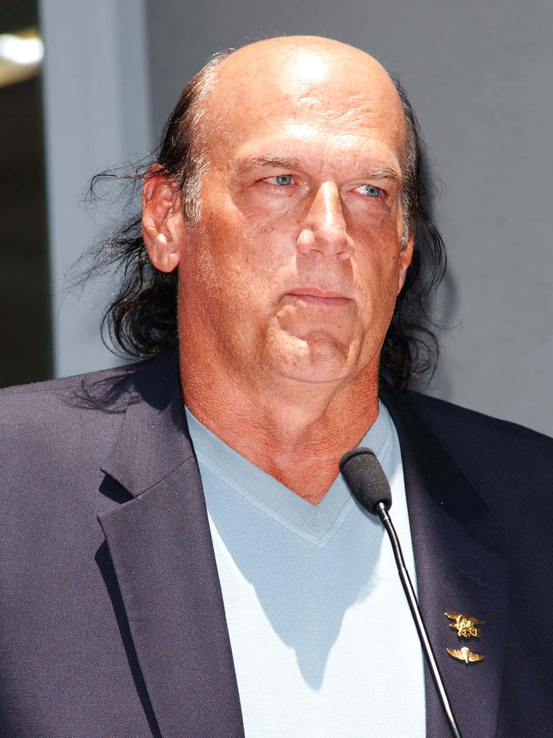明尼蘇達州州長傑西·文圖拉 壁紙 — 一位光頭長黑髮的男子穿著深色西裝和淺藍色襯衫在公共活動中的特寫照片。
