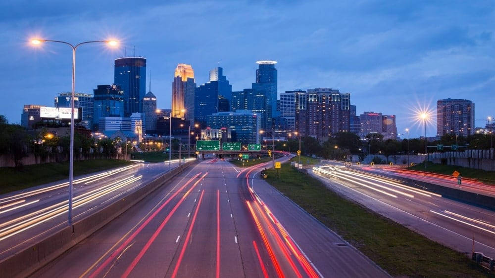 明尼阿波利斯街景夜景 壁紙 — 長時間曝光捕捉到黃昏時分高速公路上汽車燈光的軌跡，背景是閃爍的城市天際線。