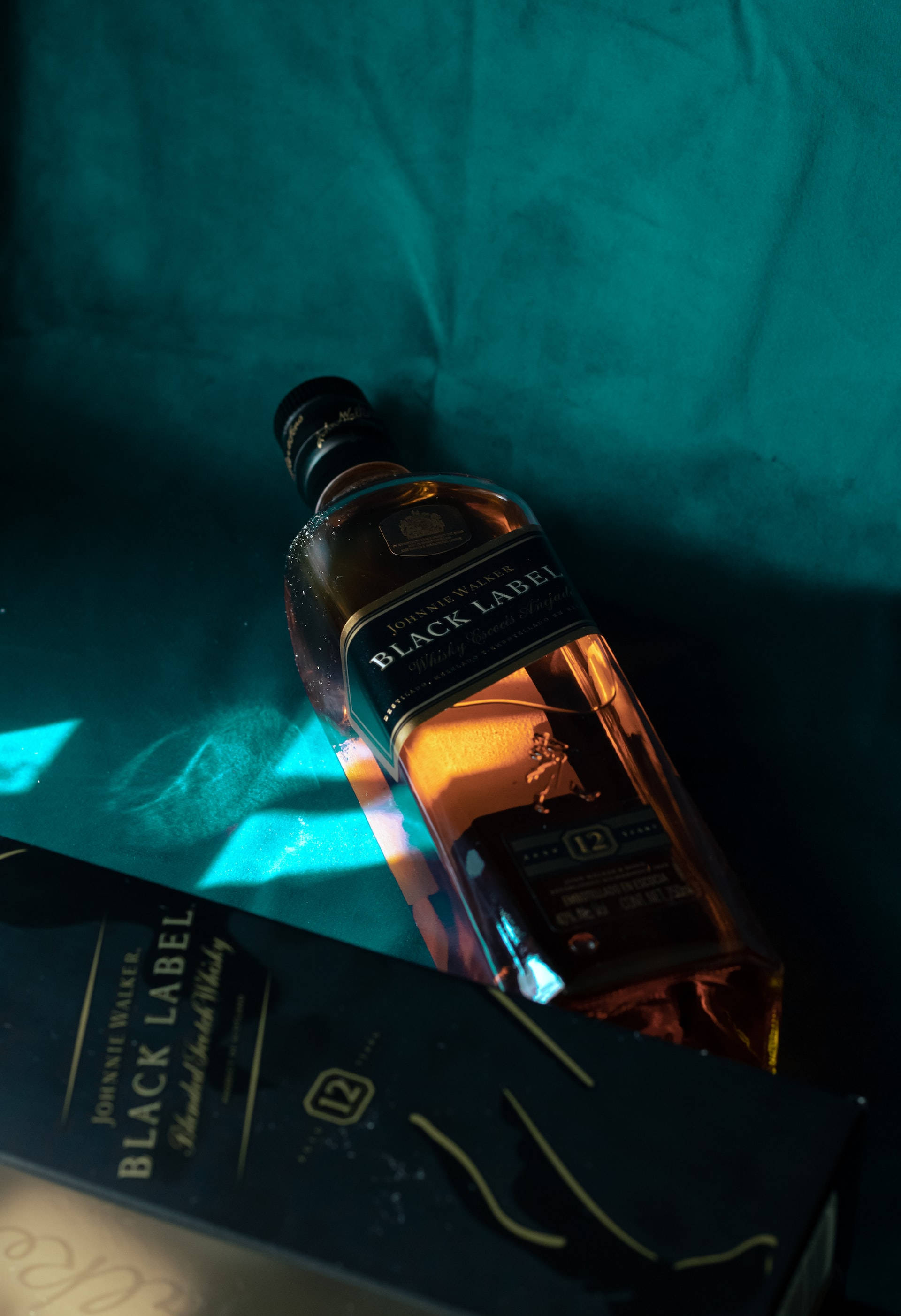 昏暗光線下的Johnnie Walker黑牌威士忌 壁紙 — 一瓶Johnnie Walker Black Label威士忌酒瓶斜放在其盒子上，背景為青綠色布料，在戲劇性的燈光下。