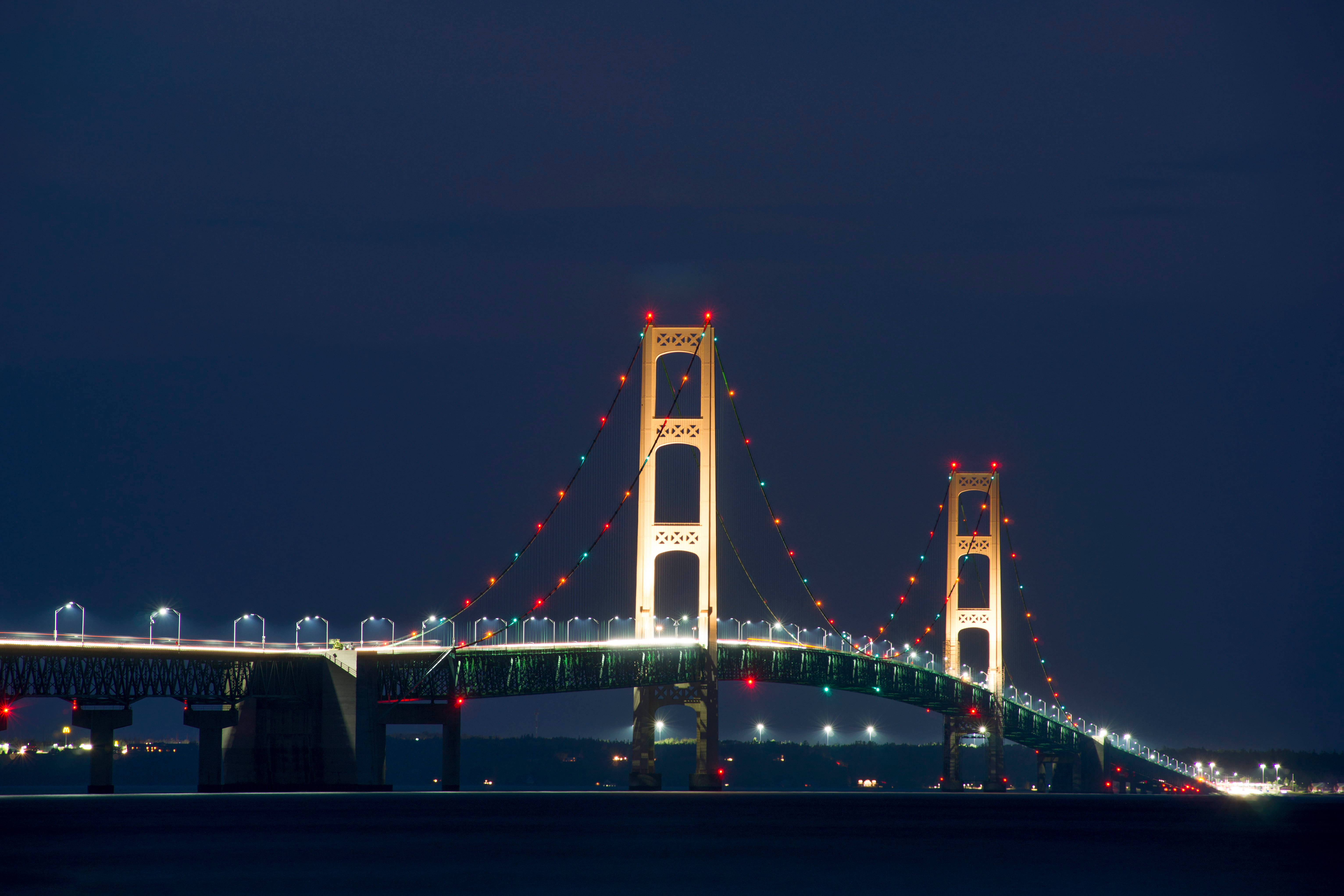 星光夜空下的燈光麥基那克橋 壁紙 — 一座宏偉的吊橋在平靜的水面上，以深沉的暮色天空為背景，閃爍著節日燈光。