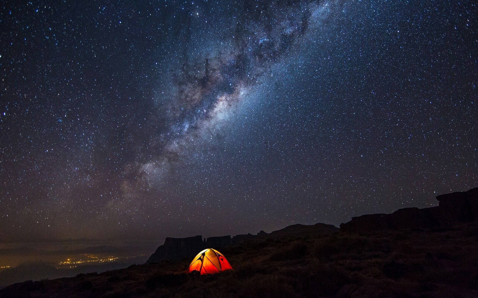 在星空下，一個發光的橙色帳篷位於山丘上，銀河清晰可見。