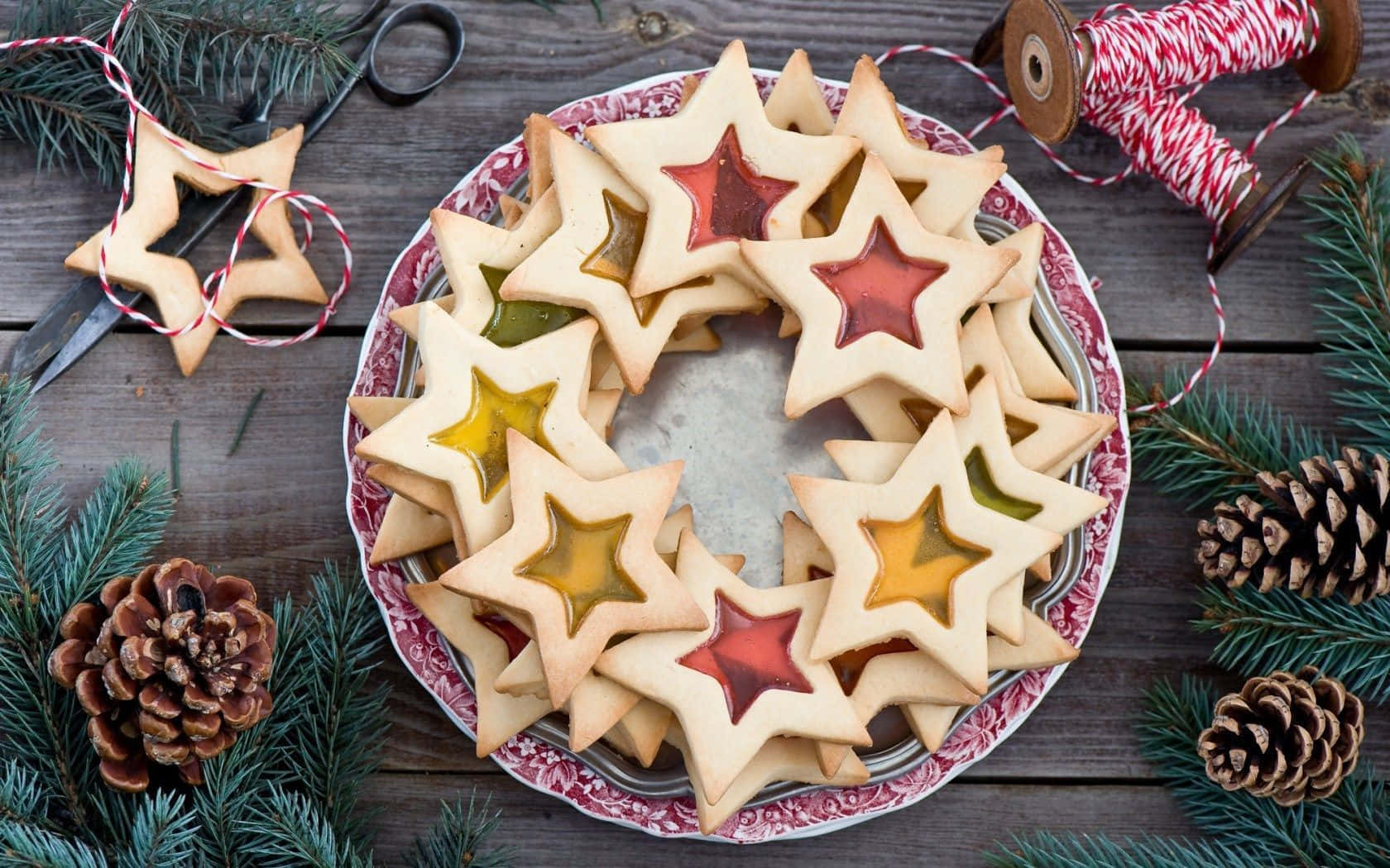 Winter Desserts 桌布 壁紙 — 星形玻璃曲奇在圖案碟上排列成花環，周圍環繞著松果和常綠樹枝。