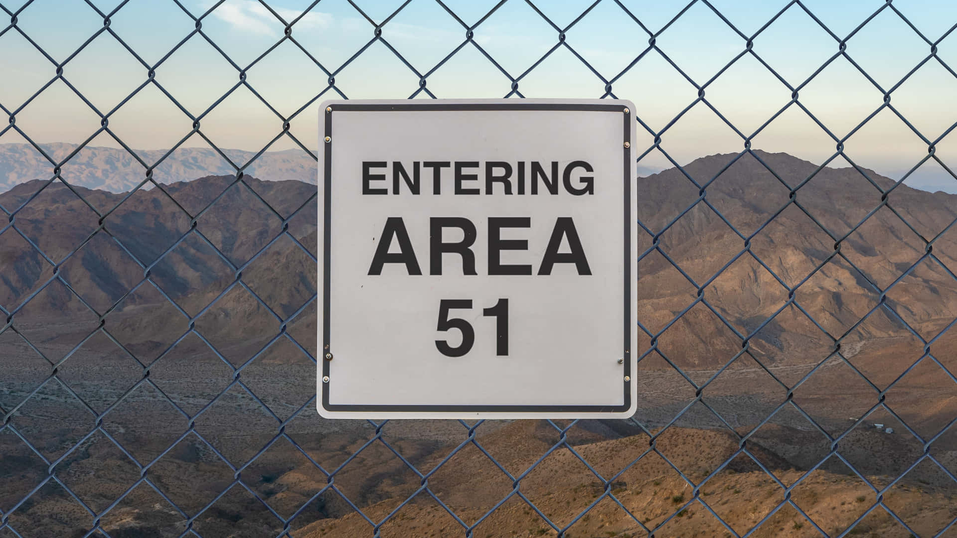 星球51 入口標誌 壁紙 — 一塊醒目的白色「ENTERING AREA 51」標誌掛在鐵絲網圍欄上，俯瞰著黃昏時分廣闊的沙漠景觀。