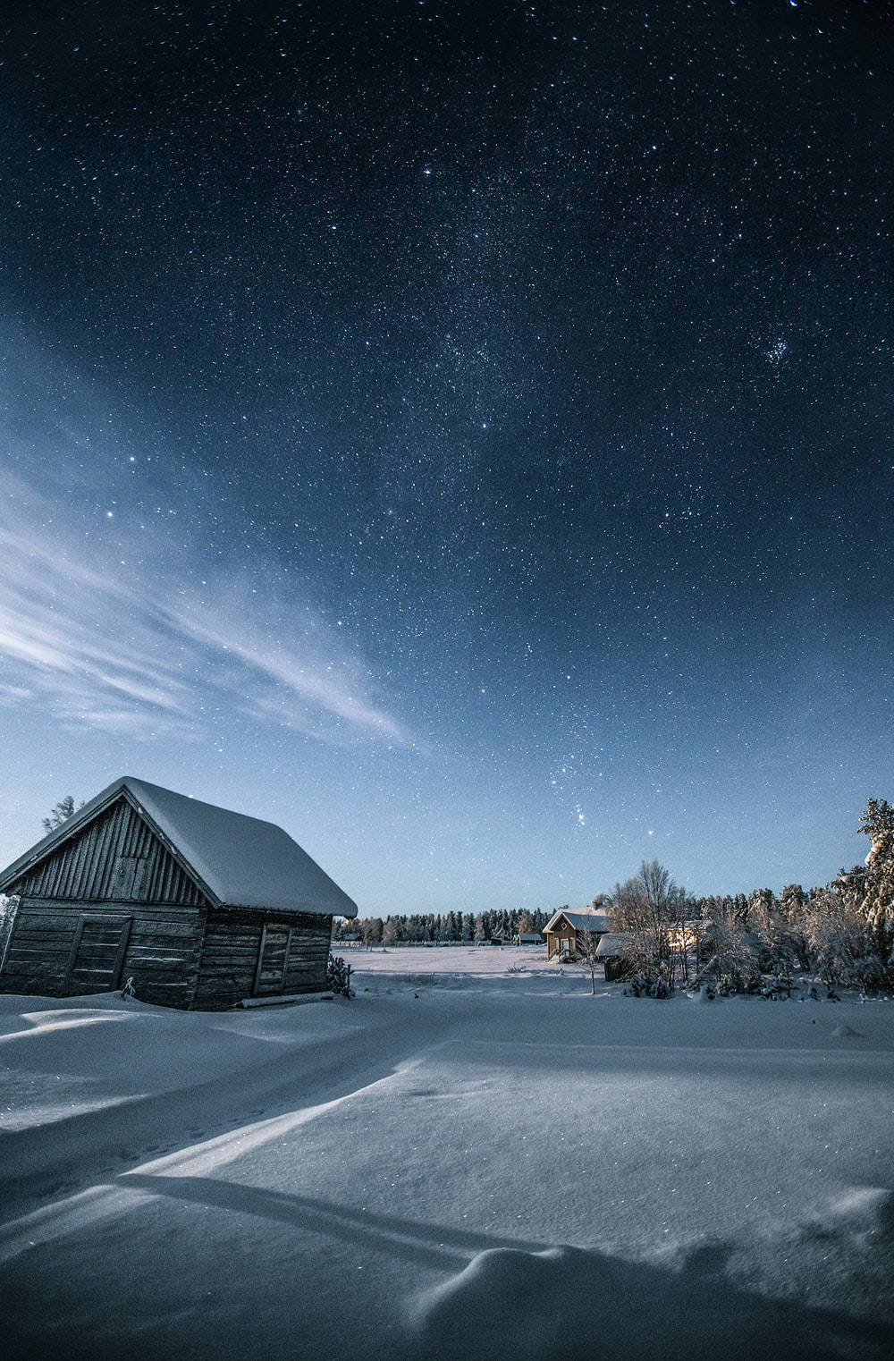 星空下的冬日小屋 壁紙 — 雪覆蓋的木屋，在繁星點點的冬夜天空下，上方有微弱的極光絲縷。