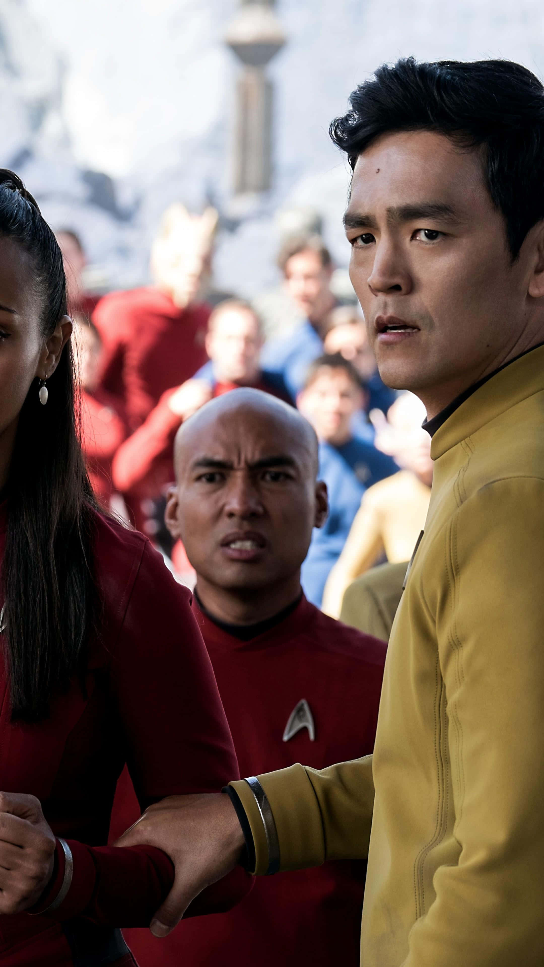 John Cho 桌布 壁紙 — 《星空奇遇記》船員穿著紅色和金色制服，在模糊的未來背景前緊張地站立。