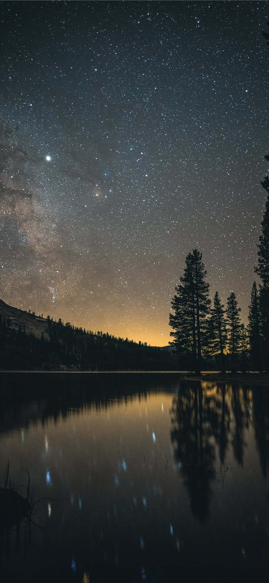 星空湖面倒影 壁紙 — 繁星點點的夜空，銀河拱起在平靜的湖面上，倒映著樹木和遠山，下方是發光的地平線。