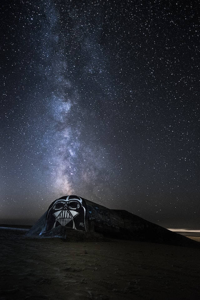 星際大戰主題iPhone壁紙 壁紙 — 一塊岩石上繪有黑武士的頭盔，在繁星點點的夜空下，銀河拱起。