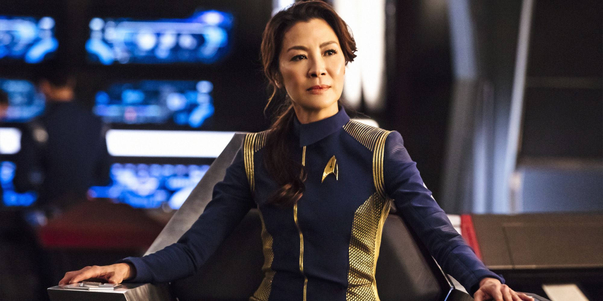 星際迷航：發現號艦長喬吉歐 壁紙 — 一名身穿藍色和金色制服嘅星際艦隊軍官，平靜咁坐喺一張未來風格嘅指揮椅上，背後有發光嘅藍色屏幕。