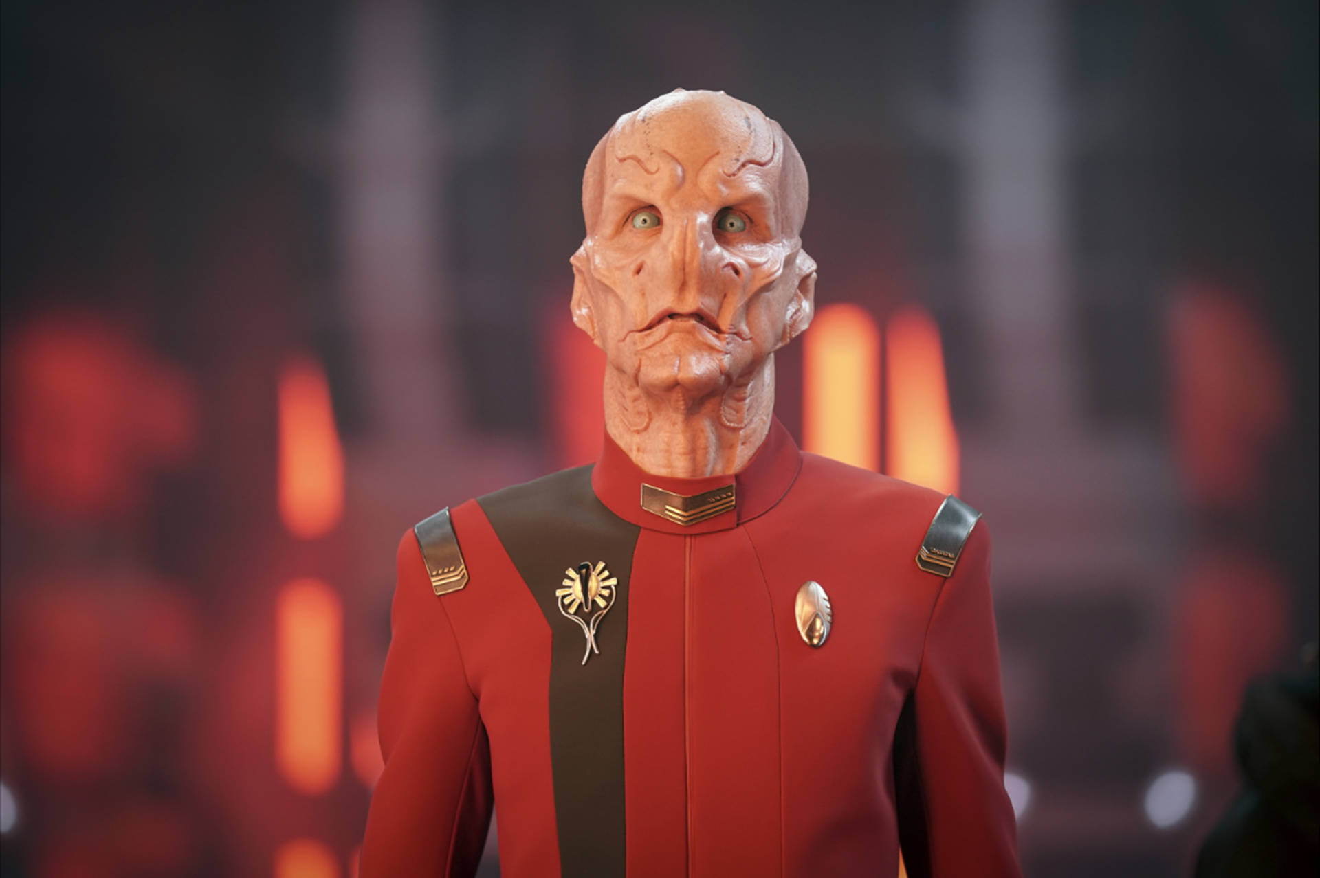 星際迷航：發現號艦長薩魯 壁紙 — 一個穿著紅色制服的人形外星人站在模糊的火焰背景前，伴隨著發光的橙色燈光。