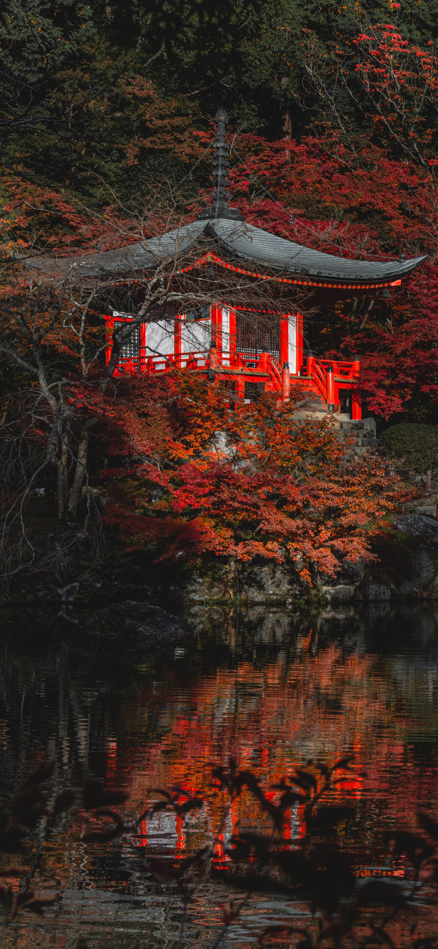 春日紅花屋 iPhone 13 Pro 壁紙 — 一座傳統的紅色日本亭閣坐落在岩石山丘上，周圍環繞著鮮豔的秋葉，其倒影在下方平靜的水面上閃爍。
