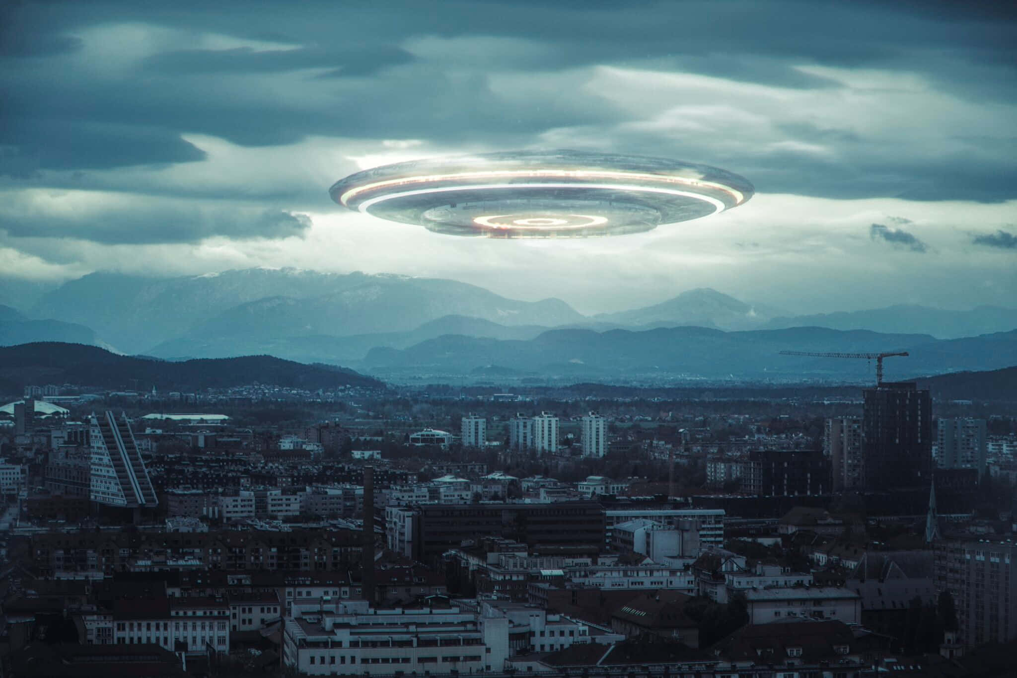 是真的！UFO確實存在！ 壁紙 — 一個發光的UFO懸浮在廣闊的城市景觀上空，背景有山脈，天空多雲。