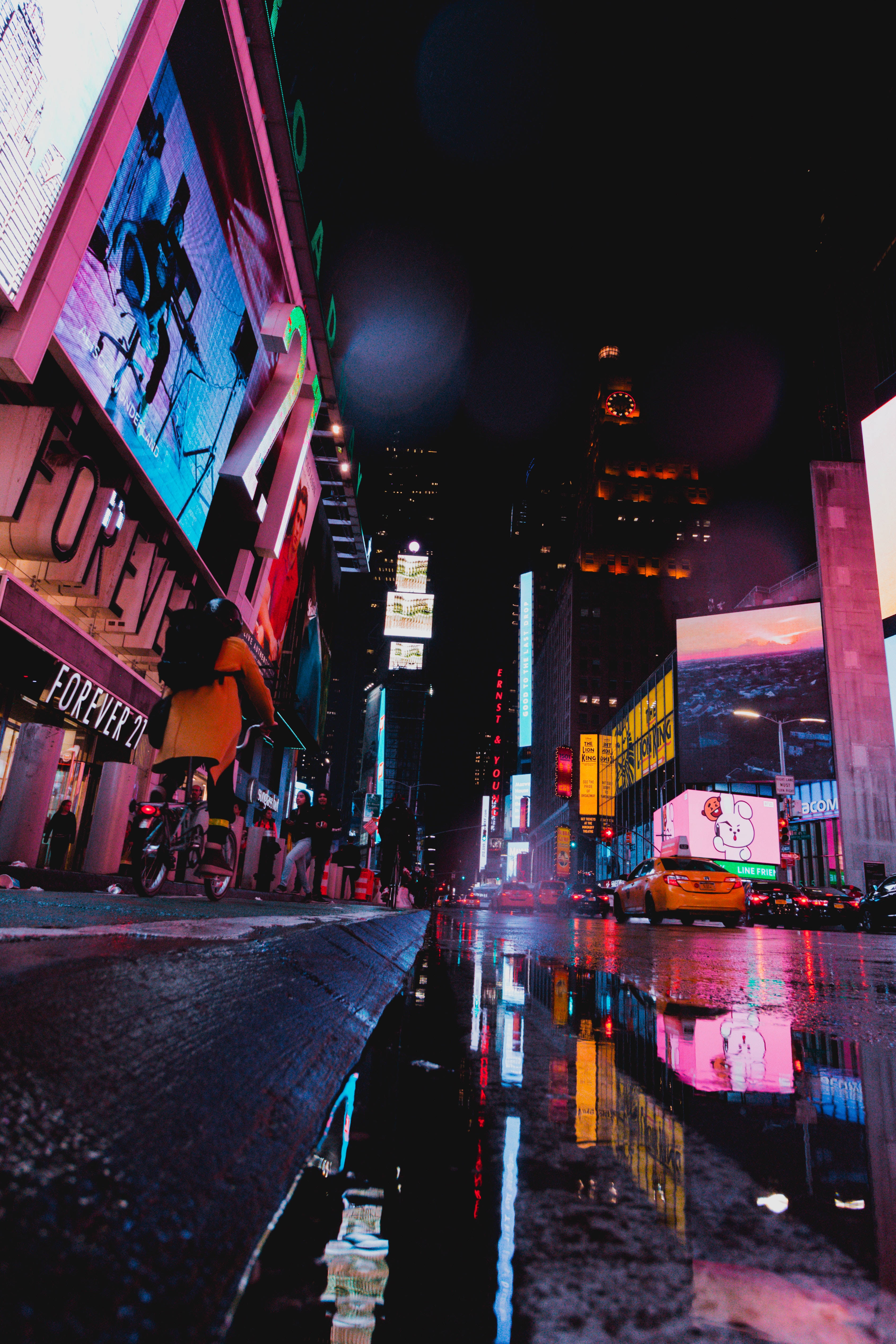 時 Square 燈光 壁紙 — 時代廣場的雨夜，霓虹燈光映照在濕漉漉的路面上，一名騎單車的人經過。