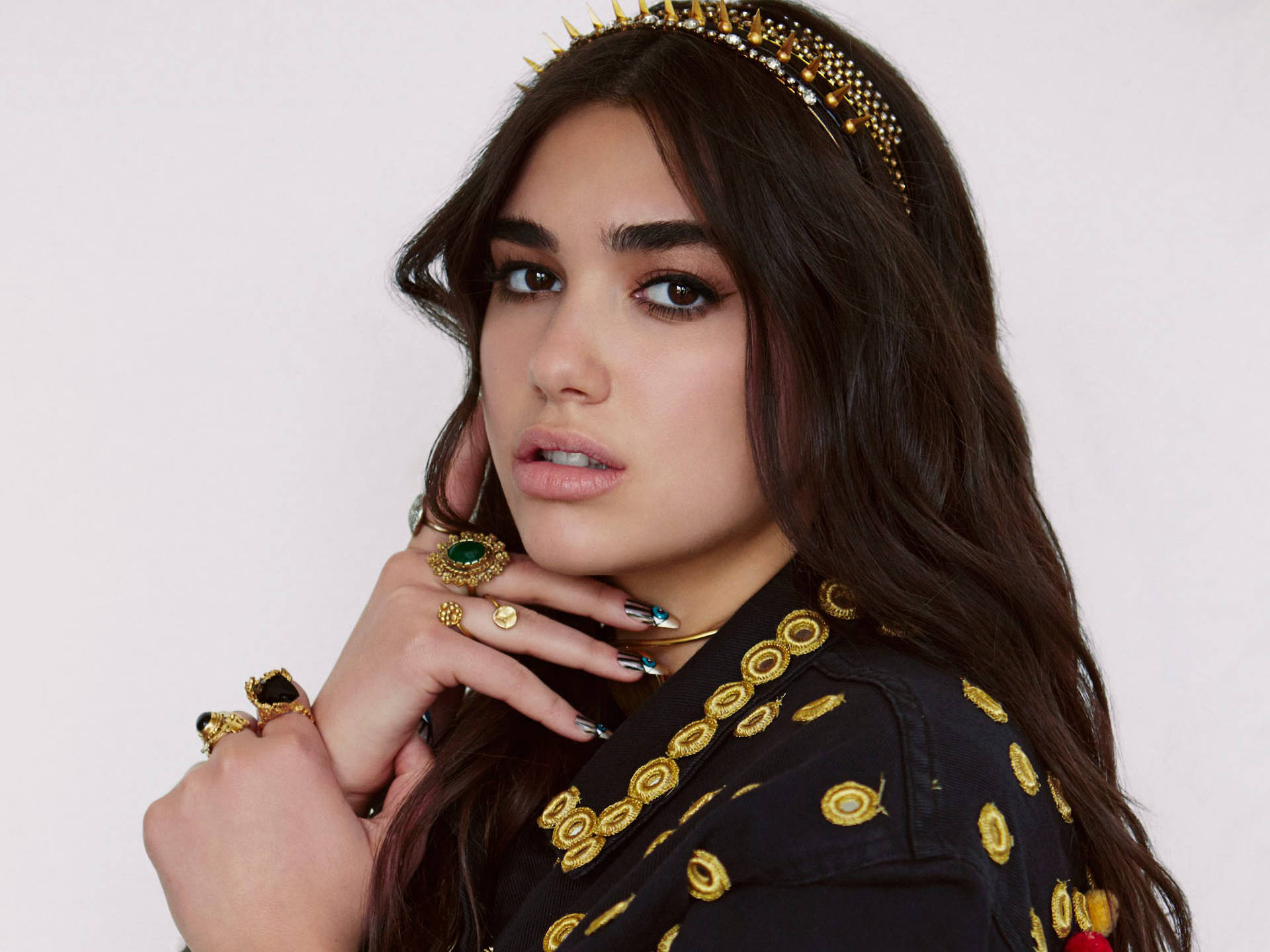 時尚 Dua Lipa 壁紙 — 一位深色頭髮的女性戴著金色尖刺頭帶和華麗戒指，在純白色背景前觸摸頸部。
