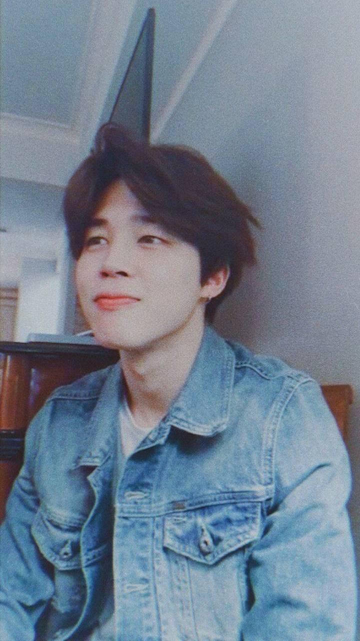 時尚 Jimin 美學 壁紙 — 一位年輕男子，棕色頭髮略顯凌亂，在柔和的燈光下室內穿著牛仔外套，露出溫柔的微笑。